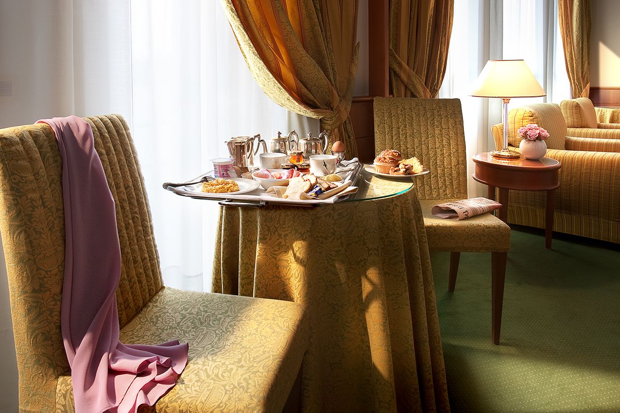В залитом солнцем гостиничном номере с драпированными шторами между двумя фактурными золотистыми стульями стоит стол, накрытый принадлежностями для завтрака.
