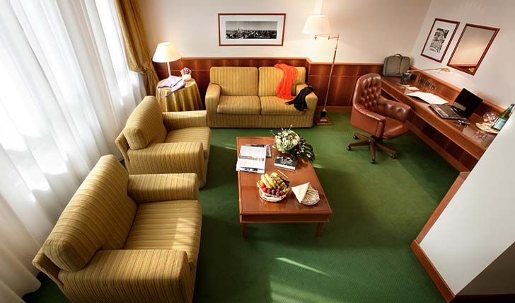 Номер в отеле с желтым диваном, двумя креслами, журнальным столиком с фруктами и письменным столом со стулом на зеленом ковре.