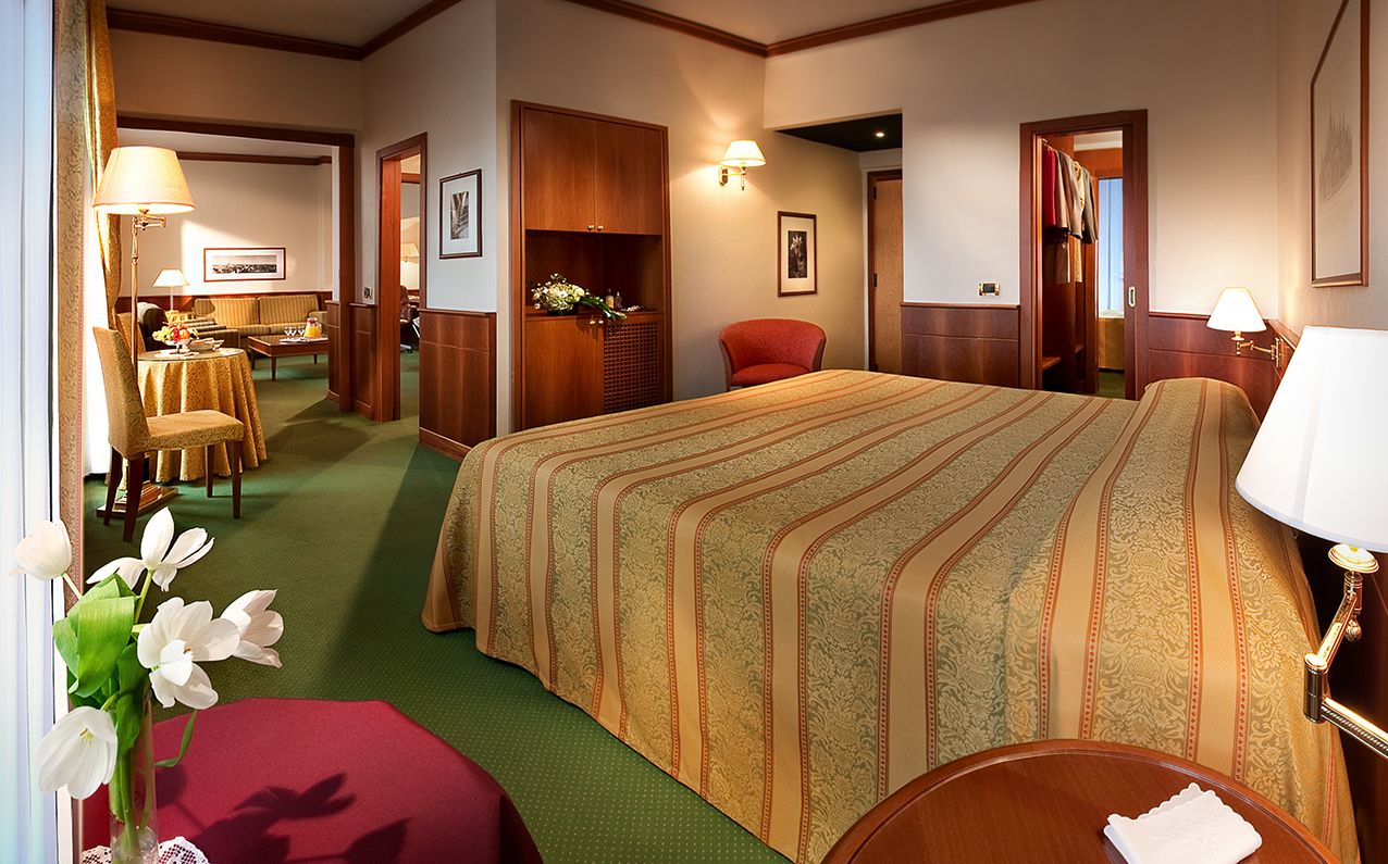 酒店房间内有一张大床，铺着金色花纹床罩，铺着绿色地毯，透过窗户可以看到隔壁的休息区。