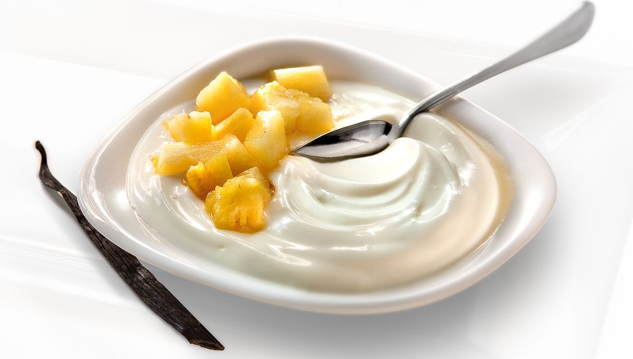 Небольшая белая миска с кремовым йогуртом, украшенная кубиками ананаса и стручком ванили, внутри которой лежит ложка.
