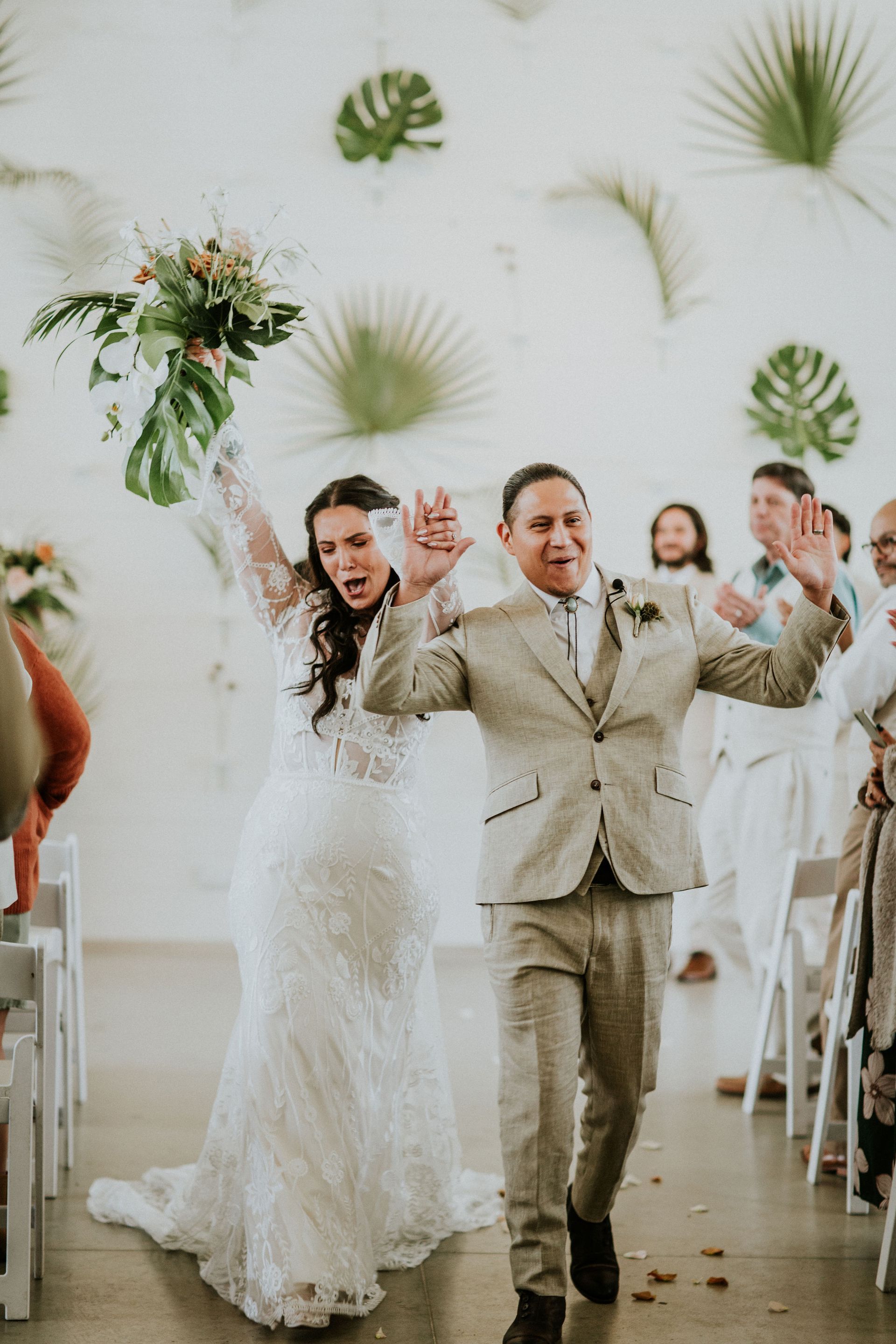 A newlywed couple celebrates while walking down an aisle, hands raised high, in front of a white wall decorated with leaves.