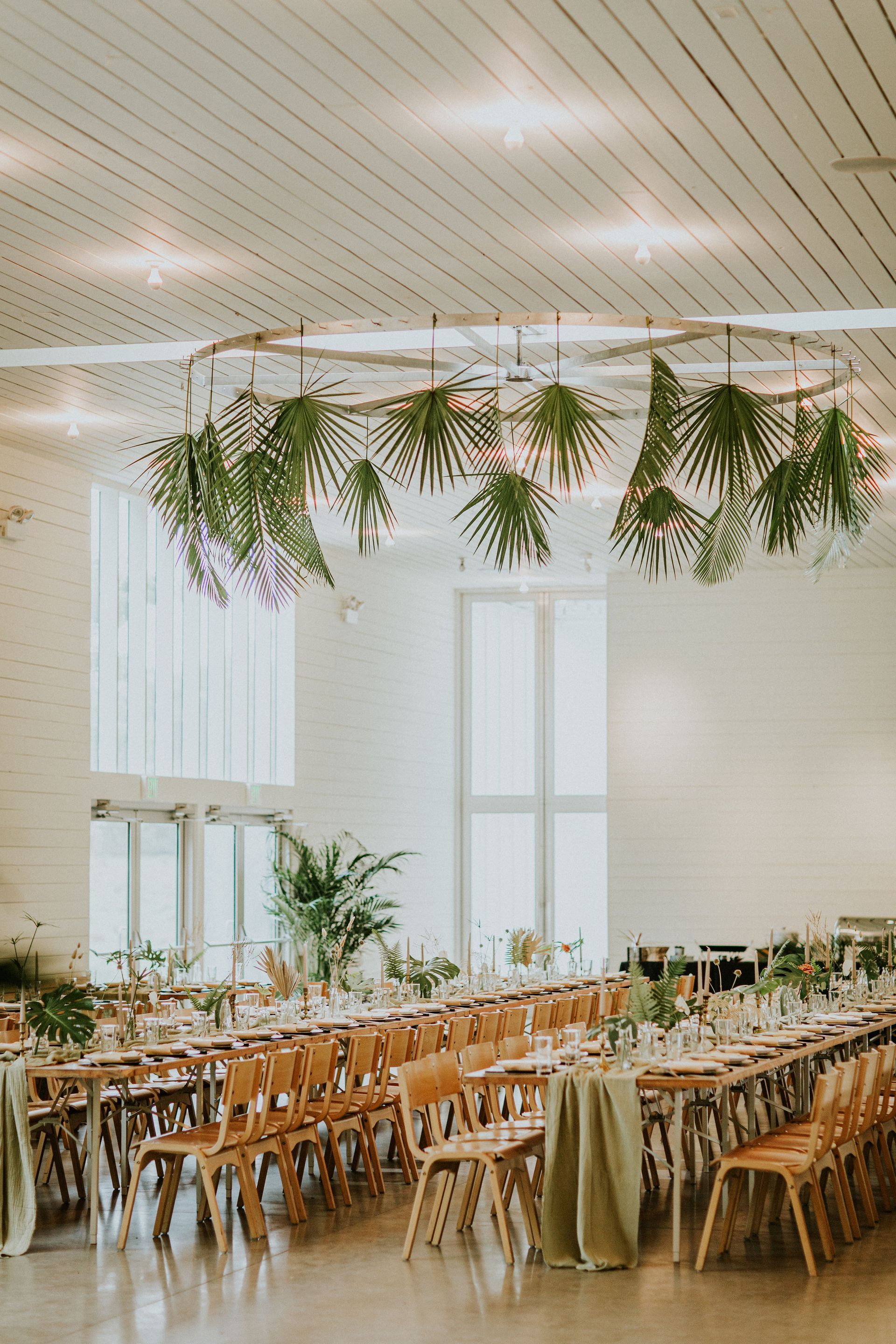 A bright event space with long rows of wooden tables, greenery-adorned chairs, and an overhead floral hanging installation.