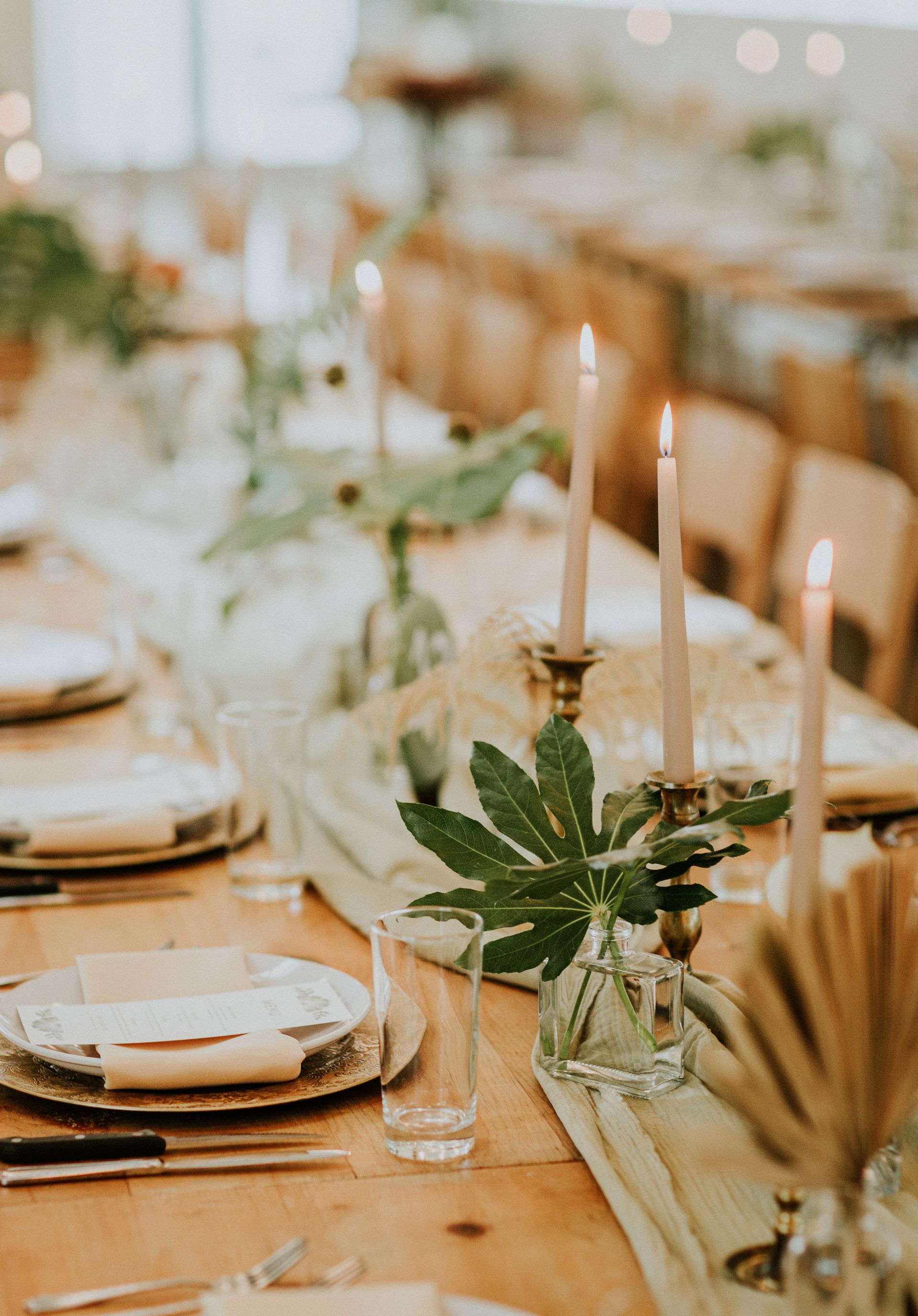 A rustic wedding table setting with taper candles, greenery in bud vases, a light green runner, and place settings.