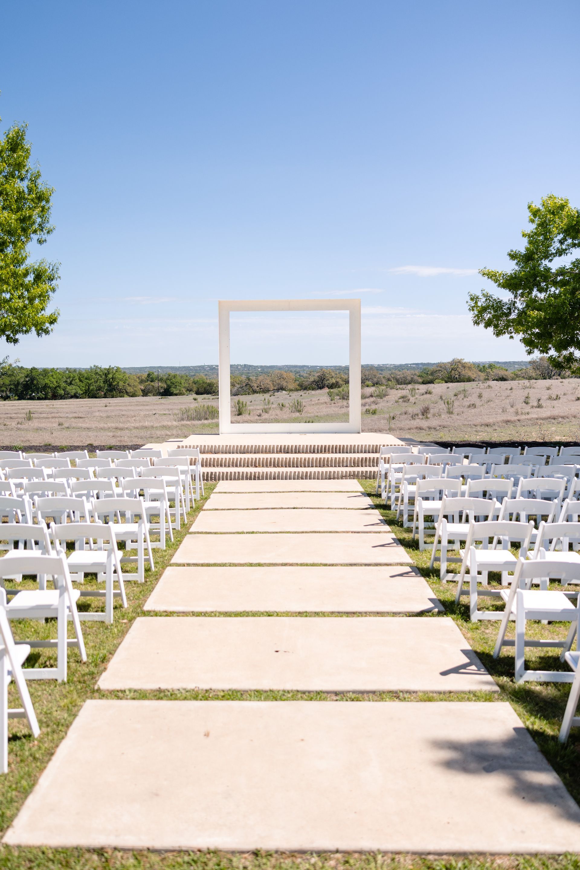 Outdoor wedding ceremony setup with rows of white chairs facing a modern, white square arch against a clear blue sky.