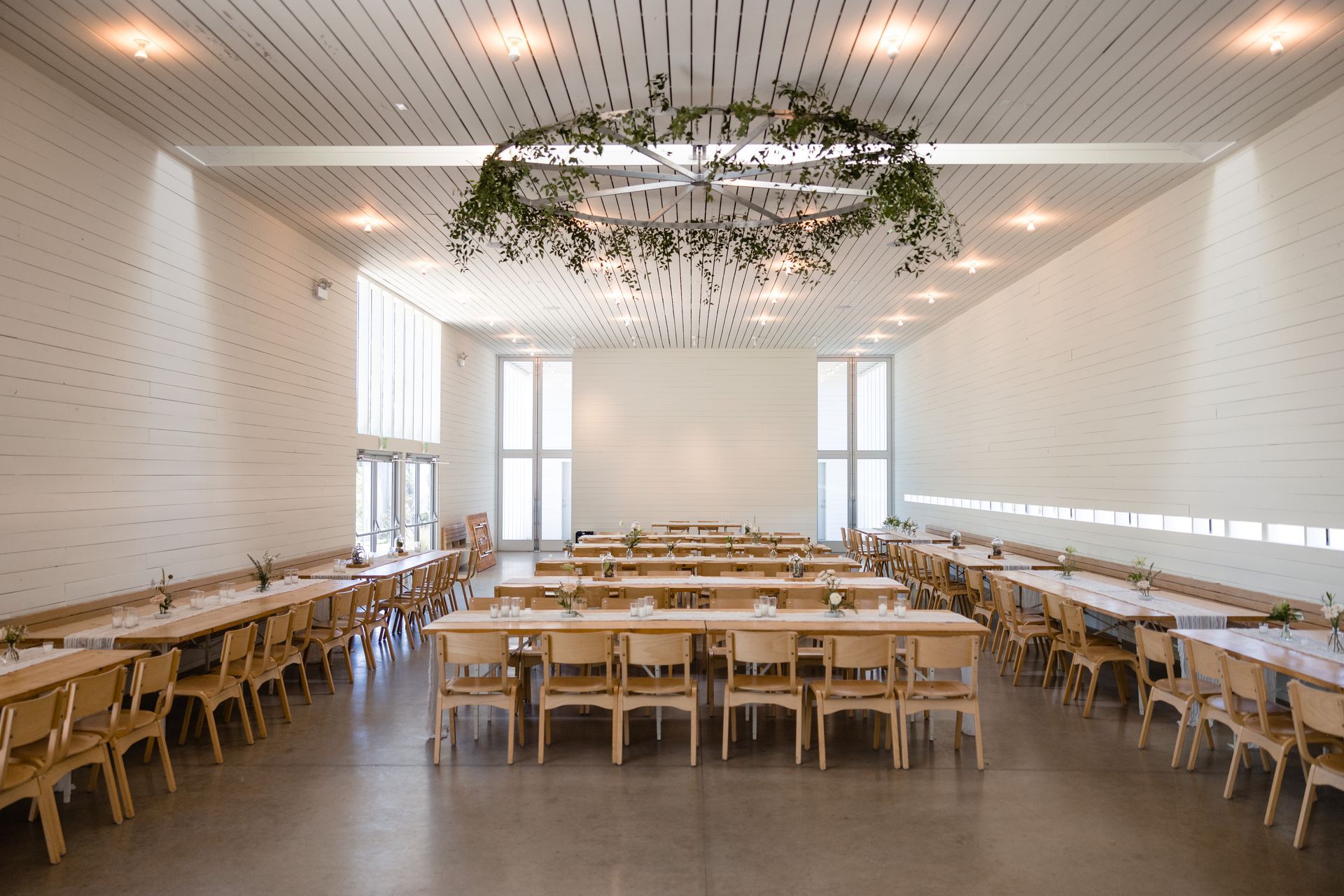 An airy, white-walled hall set for a banquet with rows of wooden tables and chairs under a large, circular greenery wreath.