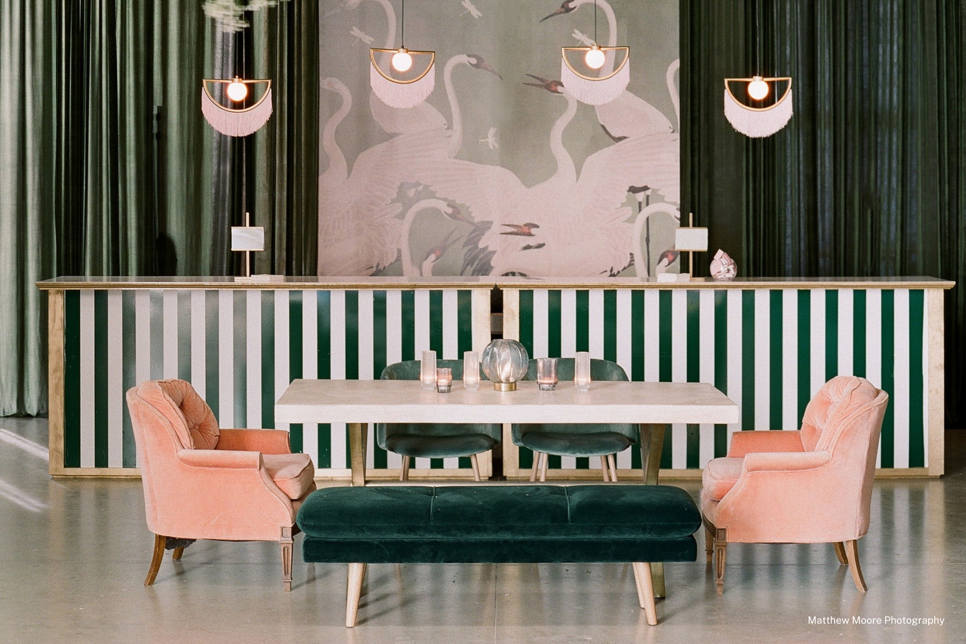 A beautiful dark green painted room with white stripes. There is a table with pink and green velvet chairs and a green velvet couch. 
