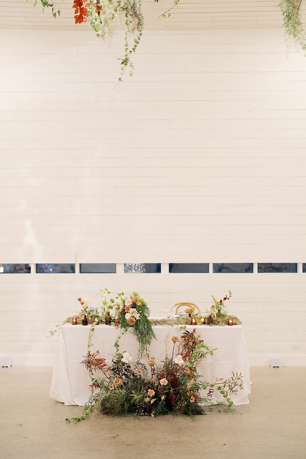 A wedding head table with a white tablecloth, decorated with lush greenery and flowers, set against a white wall at Prospect House.