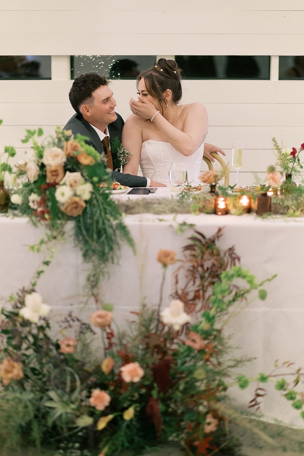 A newlywed couple sits at a floral-decorated head table, laughing and looking at each other during a reception.