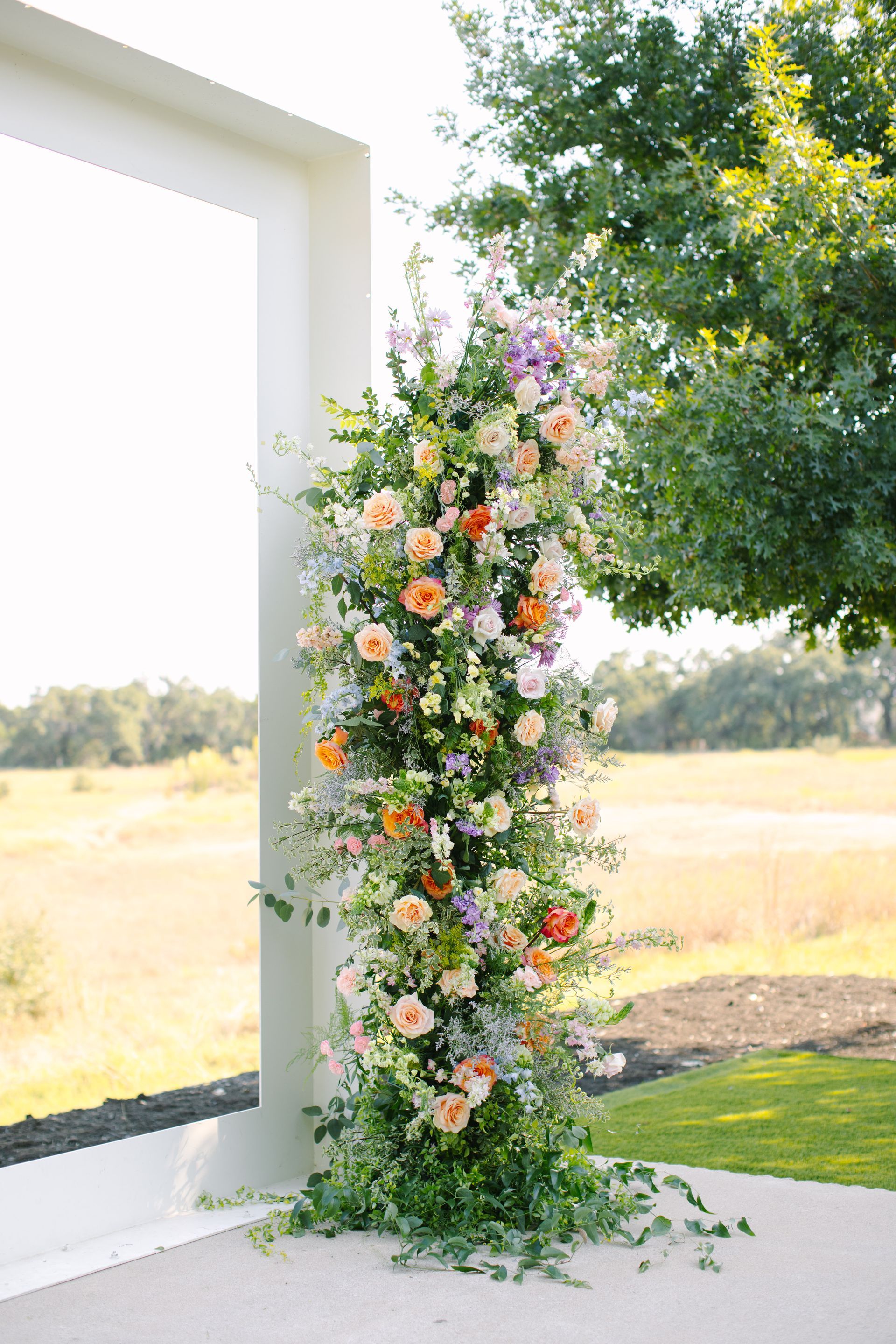A floral arrangement with orange, pink, and white blooms and greenery stands against a white frame in a grassy field.