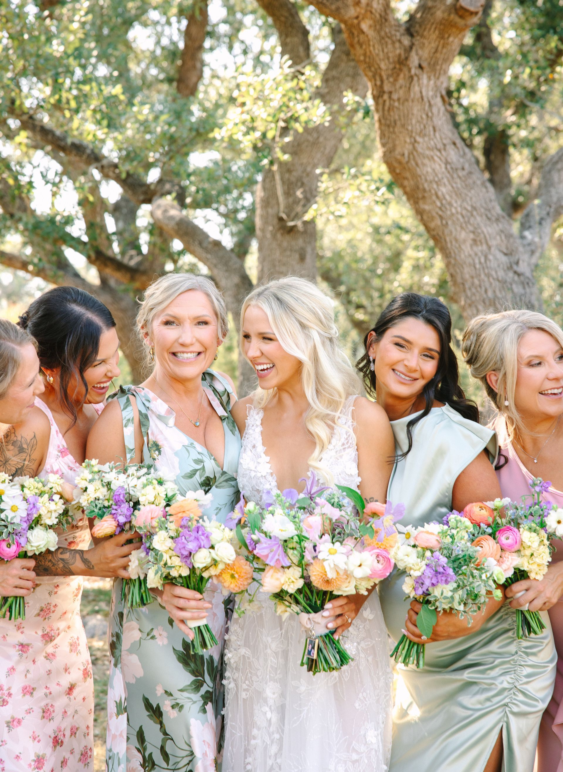 A smiling bride and five bridesmaids holding colorful bouquets, standing together outdoors in front of a tree.