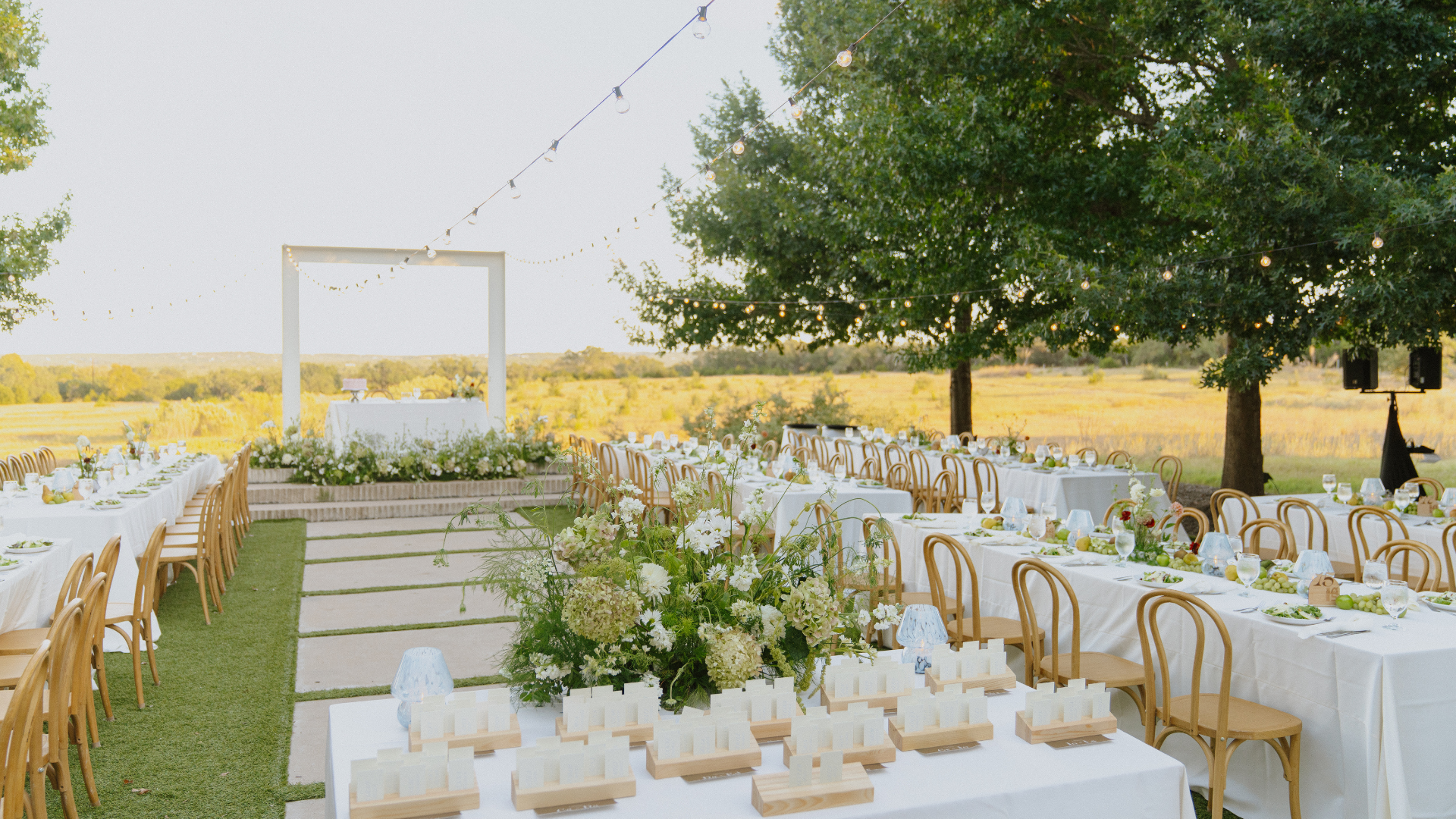 Outdoor wedding reception tables set in a garden with white flowers, gold chairs, and a white ceremony arch.