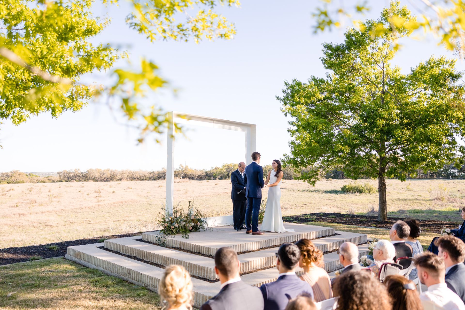 A wedding couple stands at an altar under a white arch in an open field at Prospect House, facing an officiant before a seated audience.