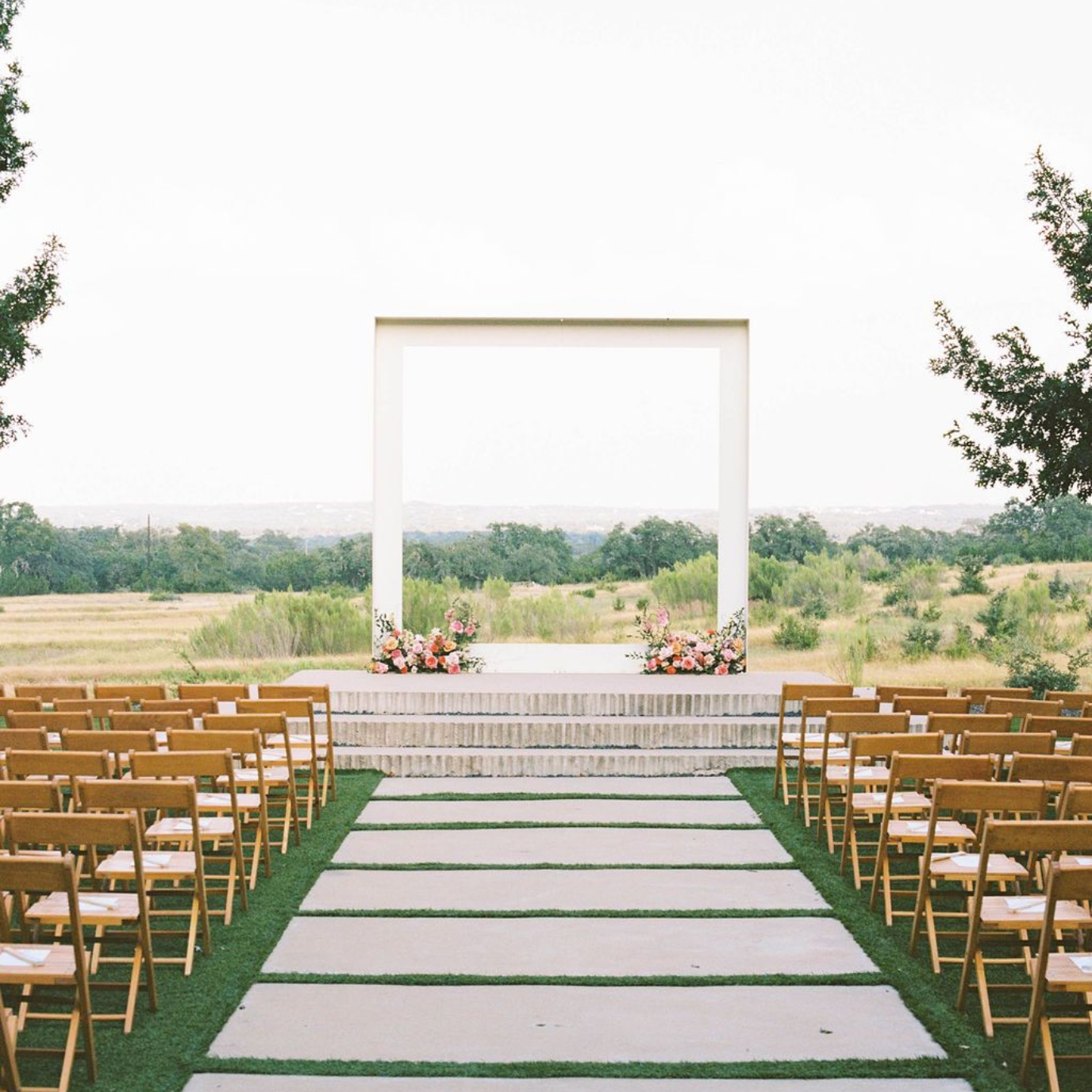 An outdoor wedding venue with rows of wooden chairs facing a white minimalist arch on a stone stage overlooking the Texas Hill Country.