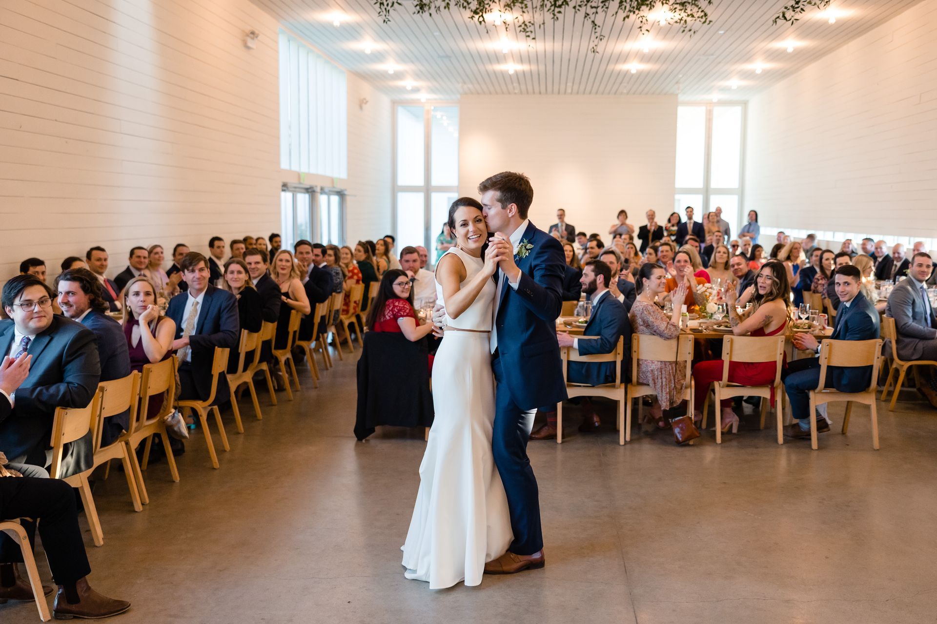 A newlywed couple dances in the center of a brightly lit event hall, surrounded by seated guests at tables.