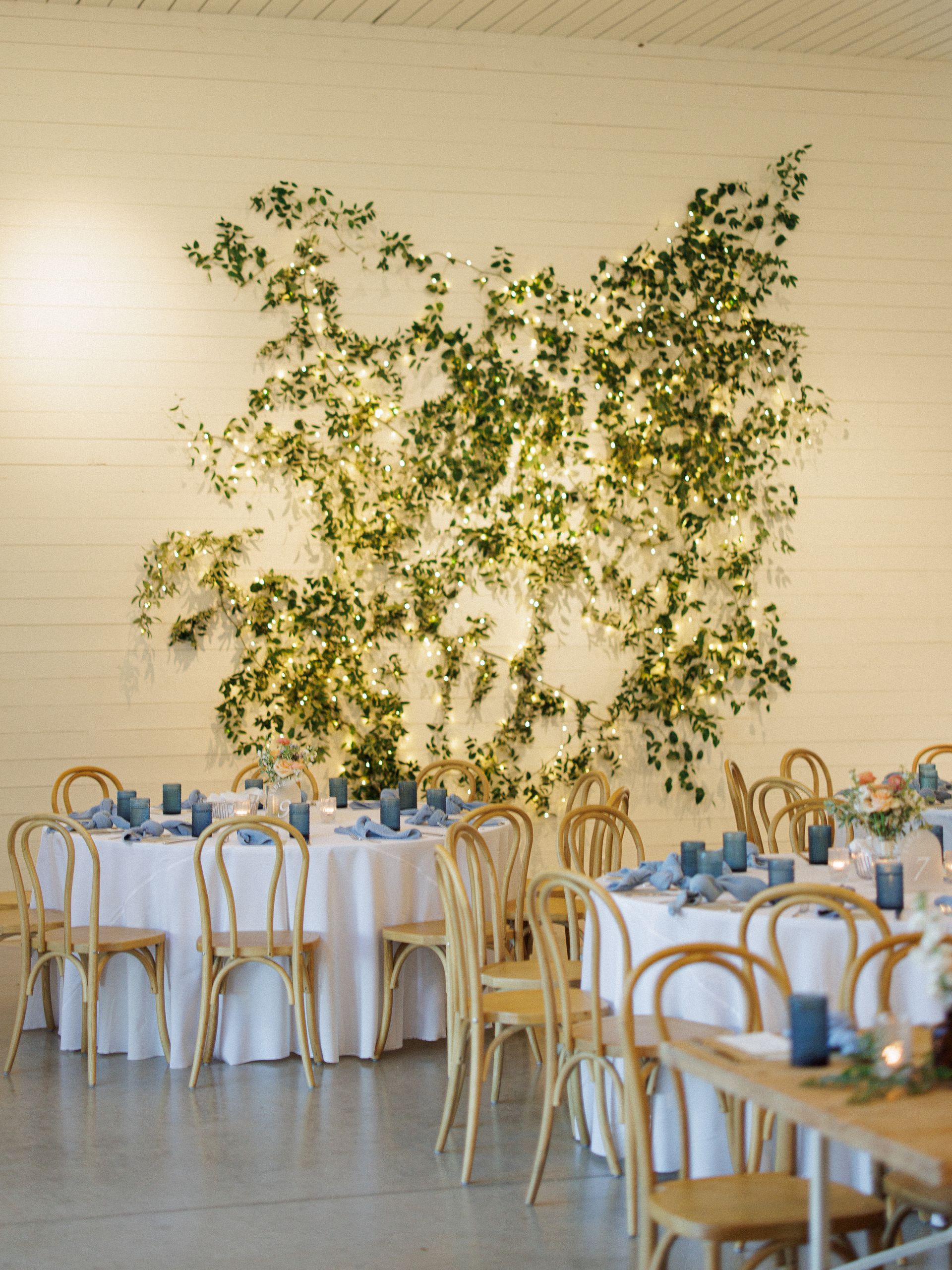 A wedding reception space with round tables, wood chairs, and a large wall decorated with lush greenery and fairy lights.