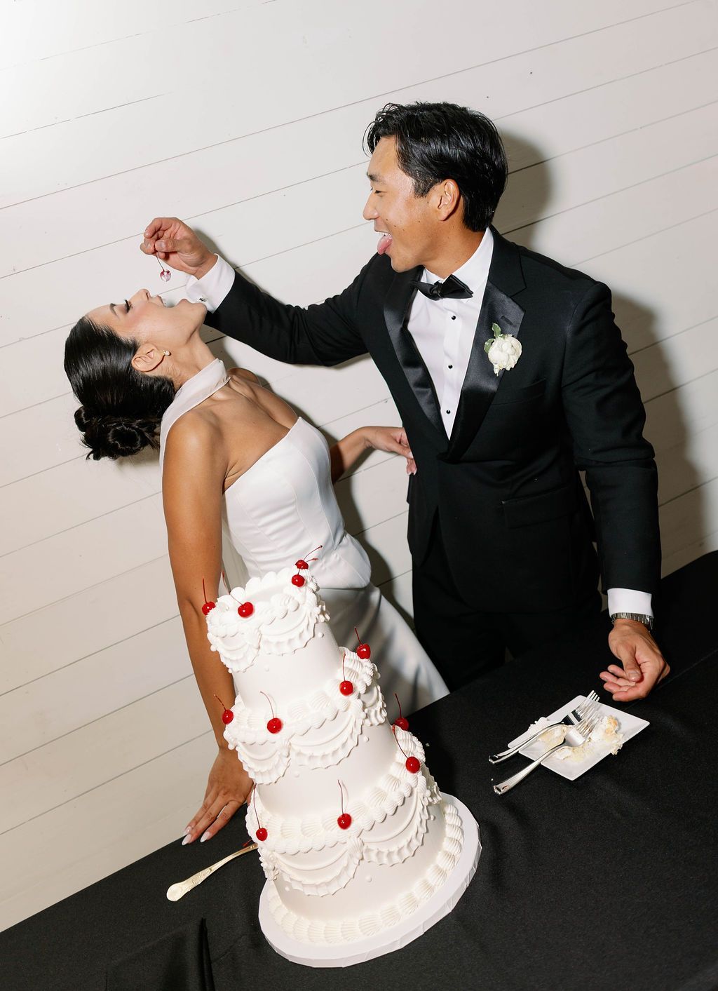 A bride and groom in formal attire playfully feed each other wedding cake in front of a white wall.
