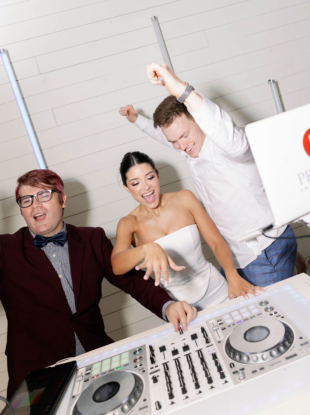 Three people celebrate while DJing at a party, one showing off an engagement ring to the camera.