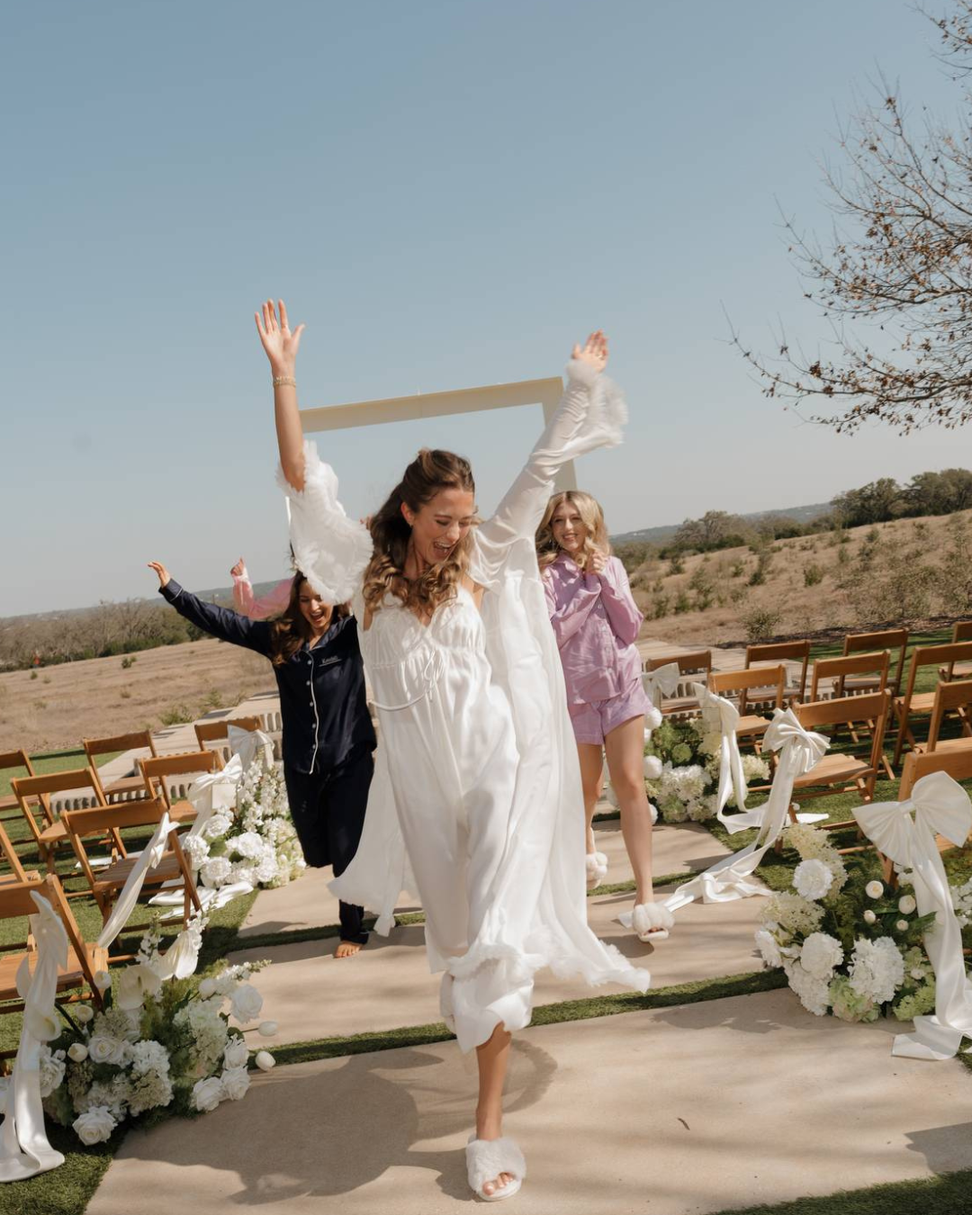 Woman in white dress dancing outdoors at a decorated wedding ceremony aisle with flowers and guests.