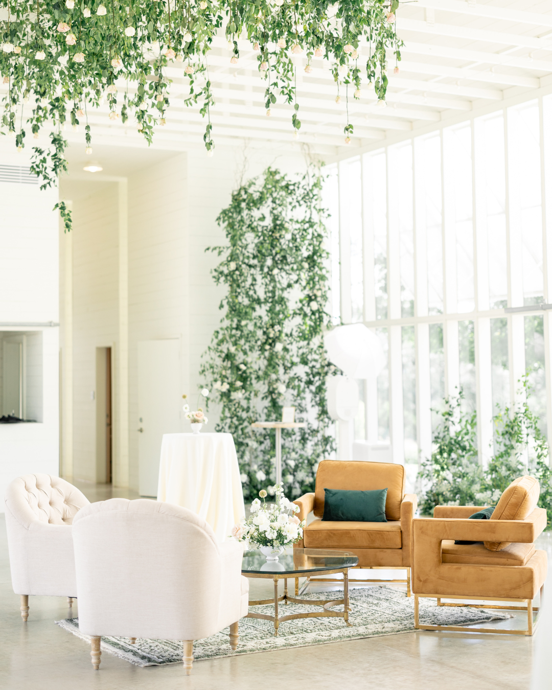 Bright sunroom with white chairs, tan sofa, and hanging green plants by tall windows