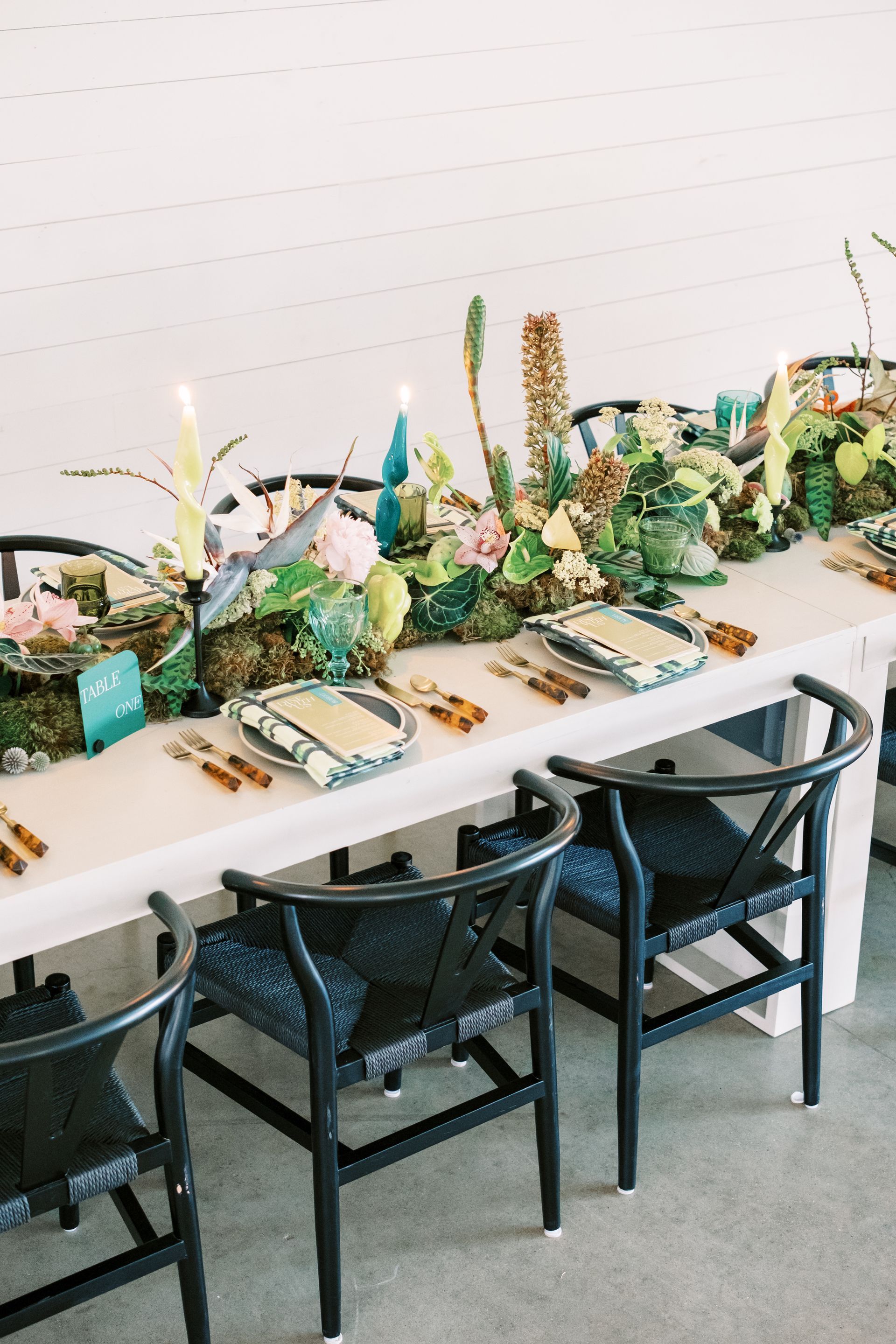A long table set with plates, utensils a menu, soft pink and green tropical flowers and candles on it.