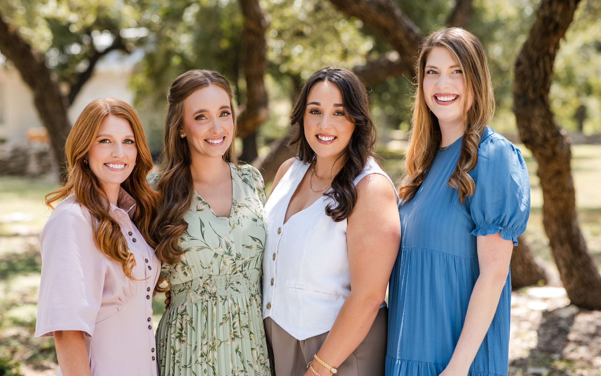 Four smiling women stand side-by-side outdoors in front of trees, wearing colorful casual dresses and a vest.