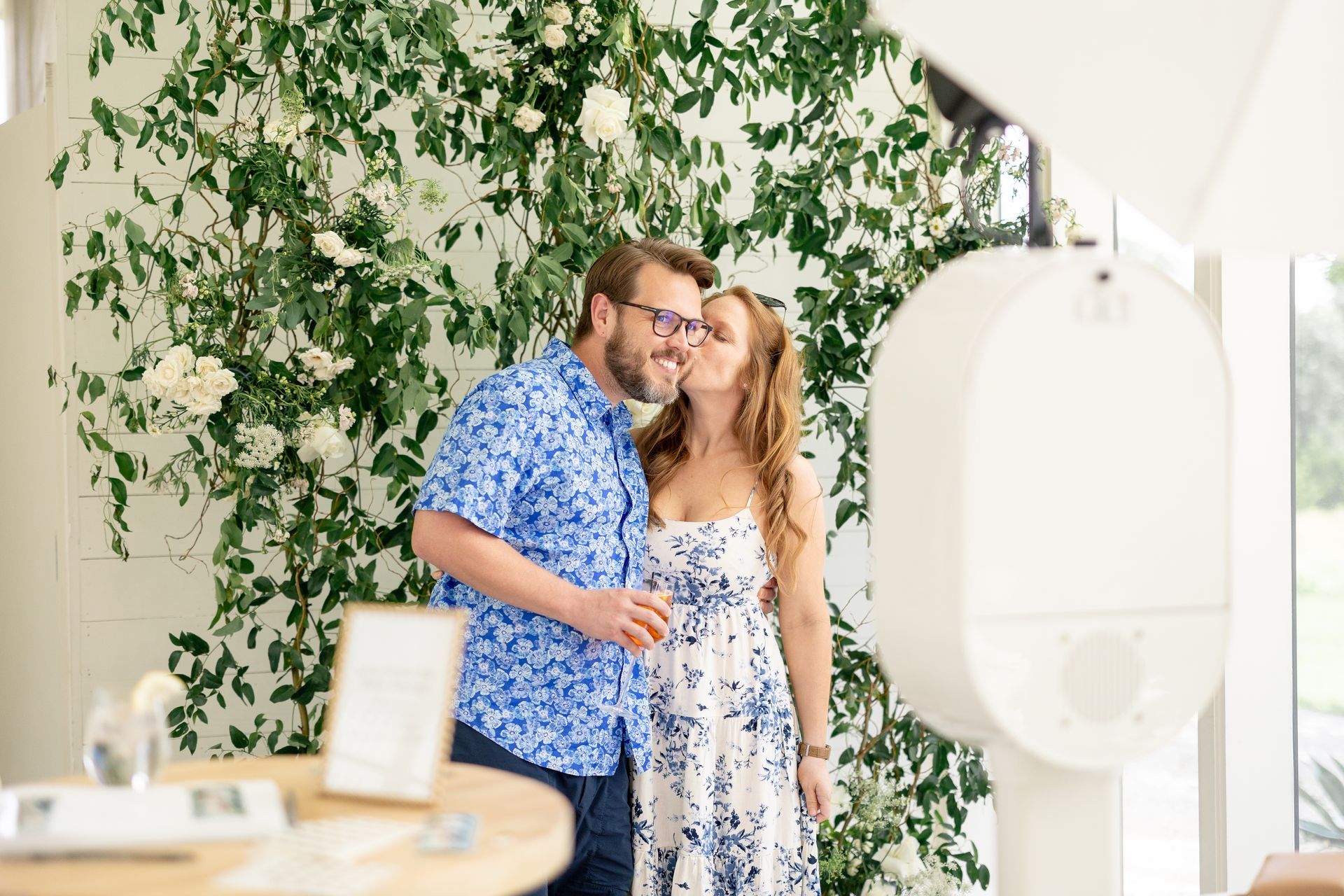 A woman in a blue and white floral dress kisses a man in a matching floral shirt in front of a floral photo booth.