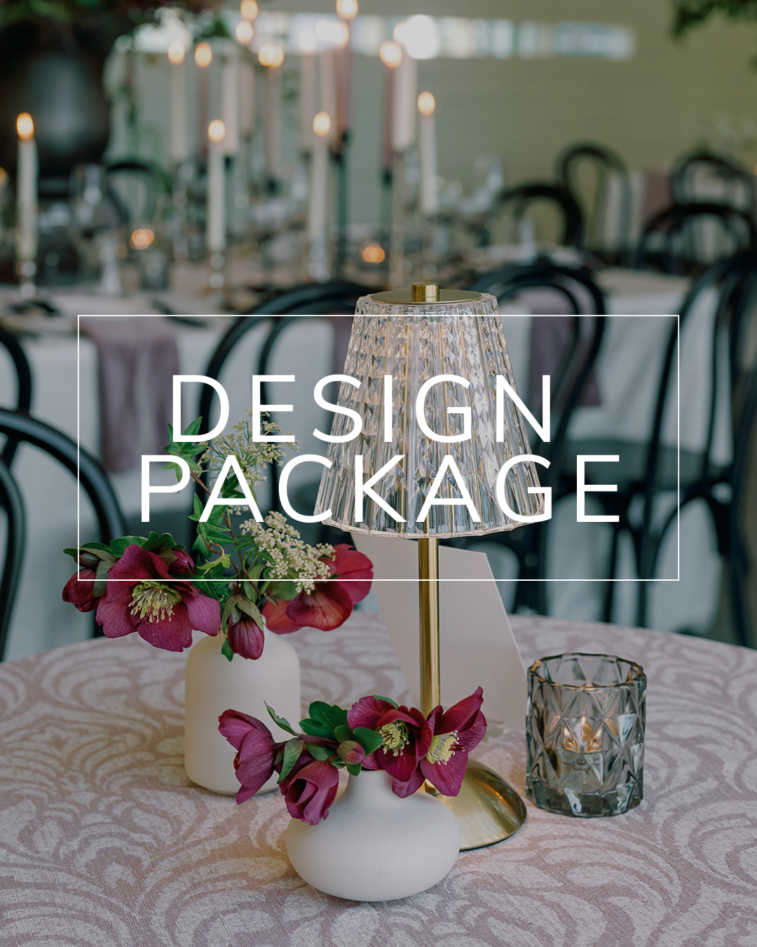 A wedding table centerpiece with mauve flowers, two white vases, a gold-stemmed lamp, and a candle on a patterned cloth.