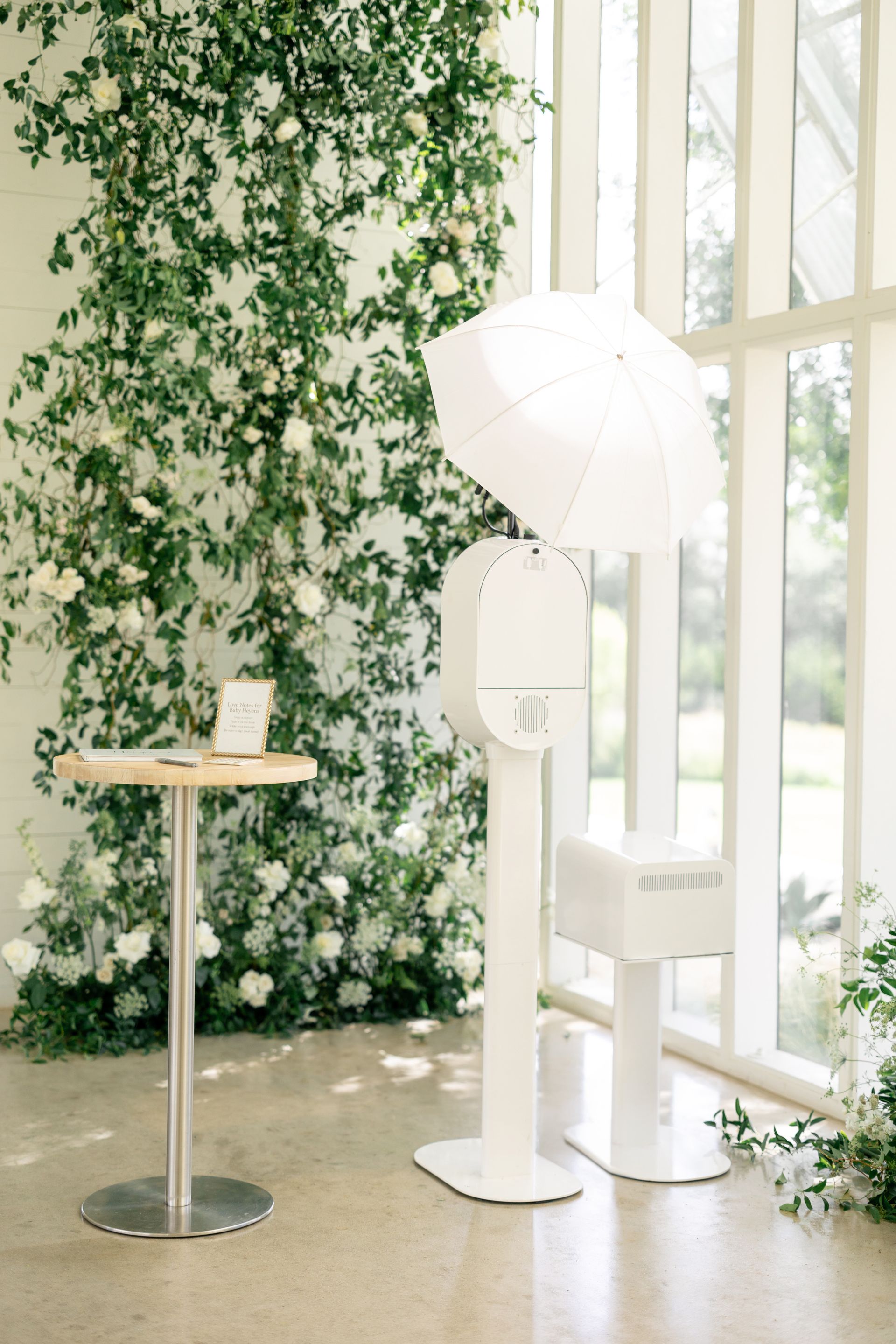A white photo booth with an umbrella and a matching table set against a wall adorned with greenery and white roses.