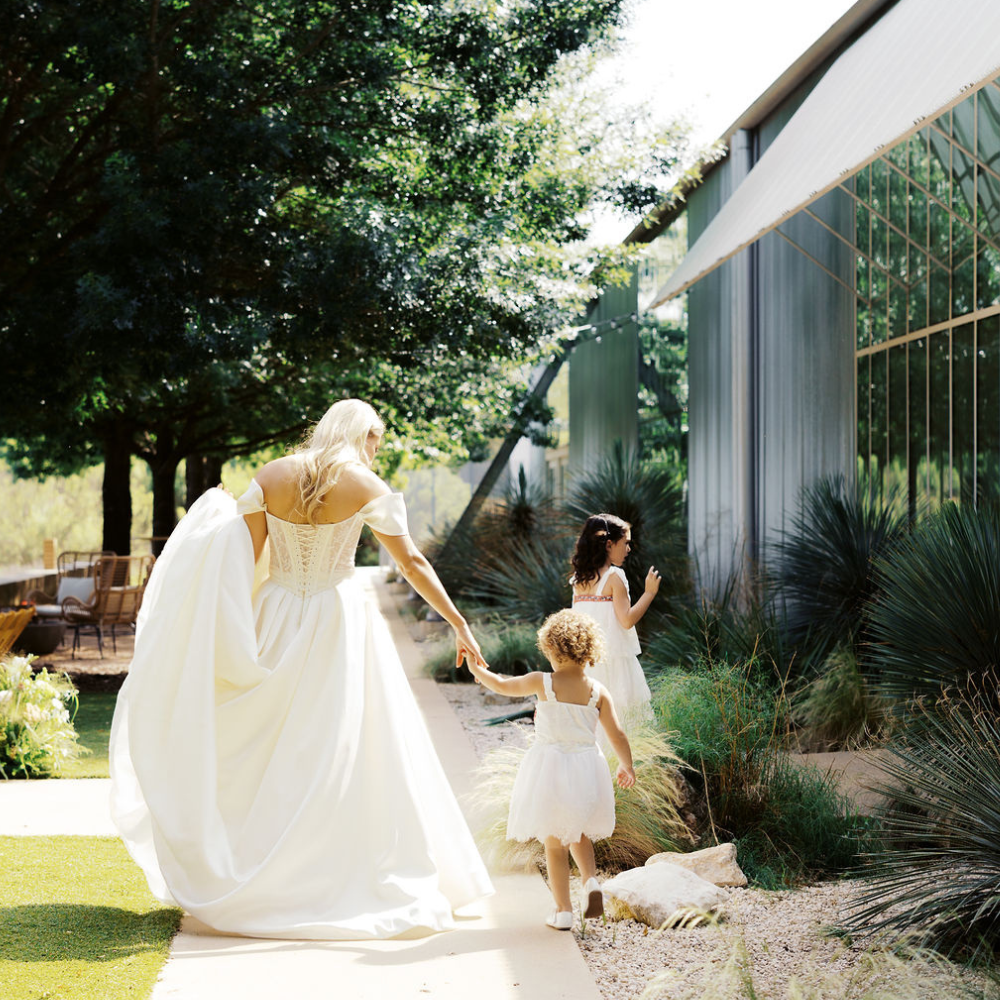 Outdoor wedding venue with a paved path, white patio umbrellas, and guests mingling near a white structure under trees.