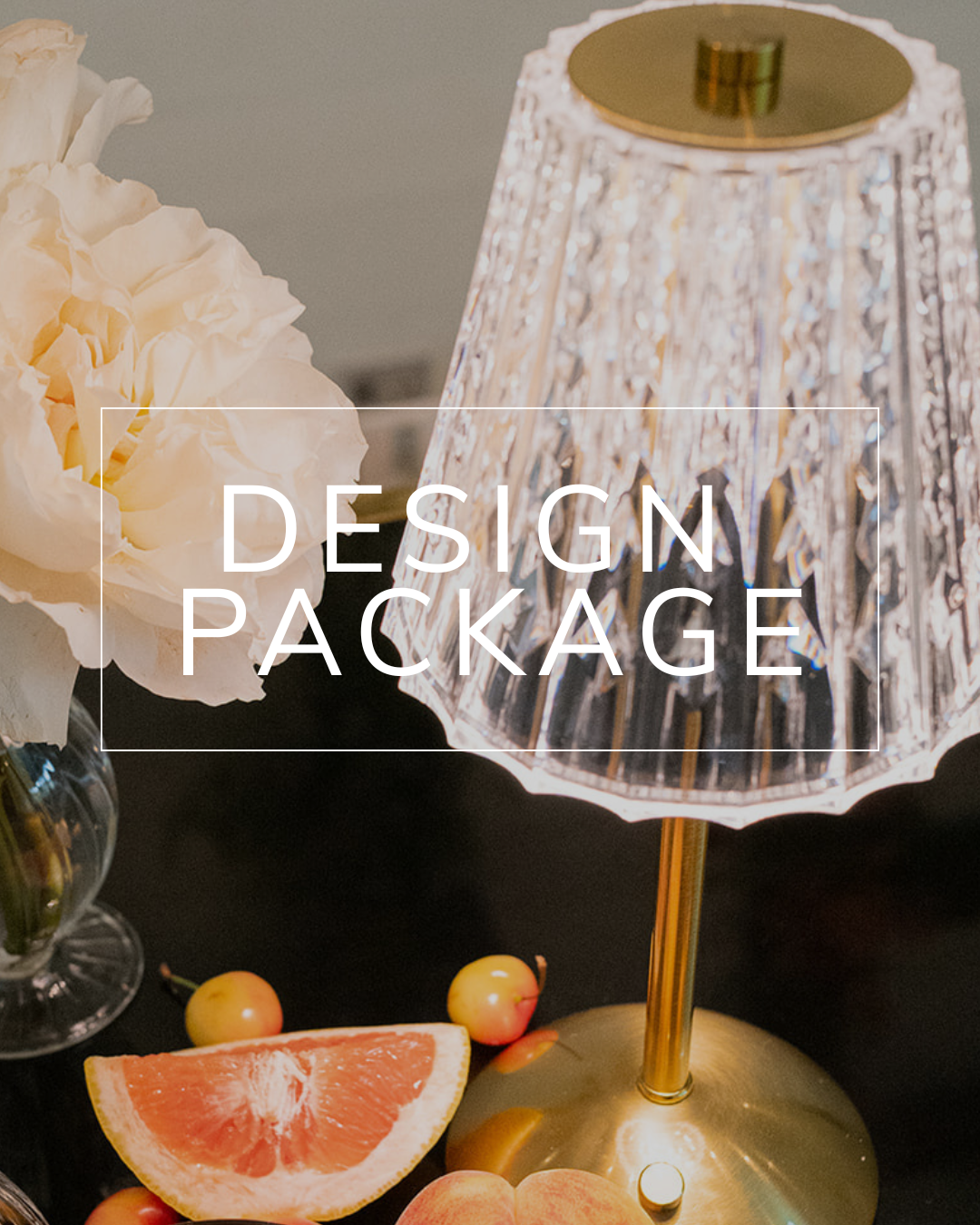 A gold and glass table lamp sits next to a vase with white flowers and fruit on a dark table, with