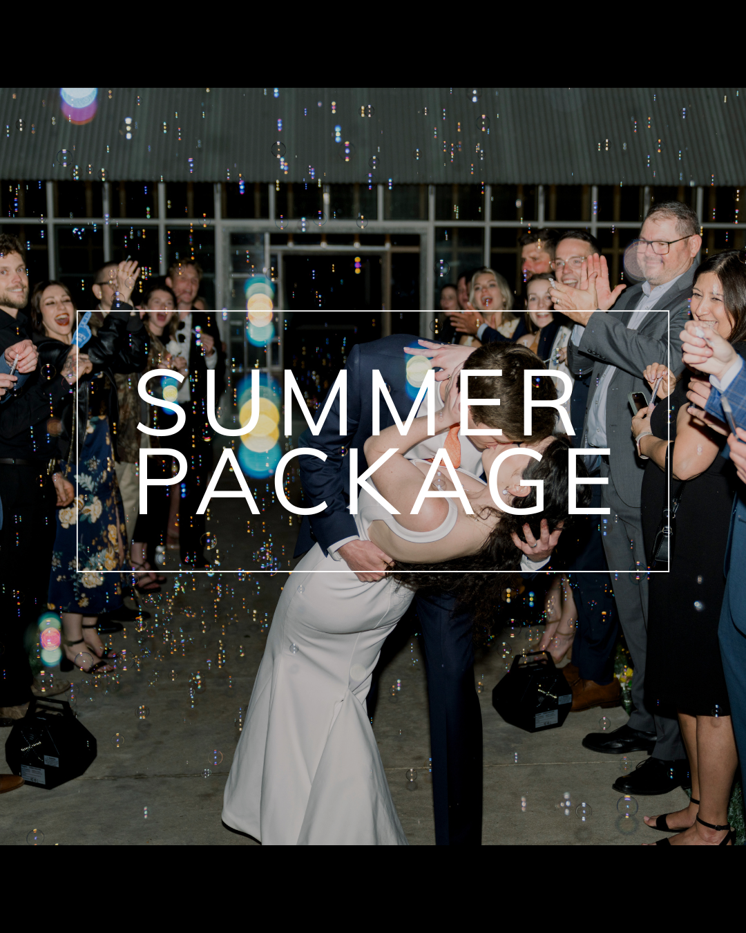 A couple kisses as they are showered with bubbles, surrounded by wedding guests at an evening outdoor reception.