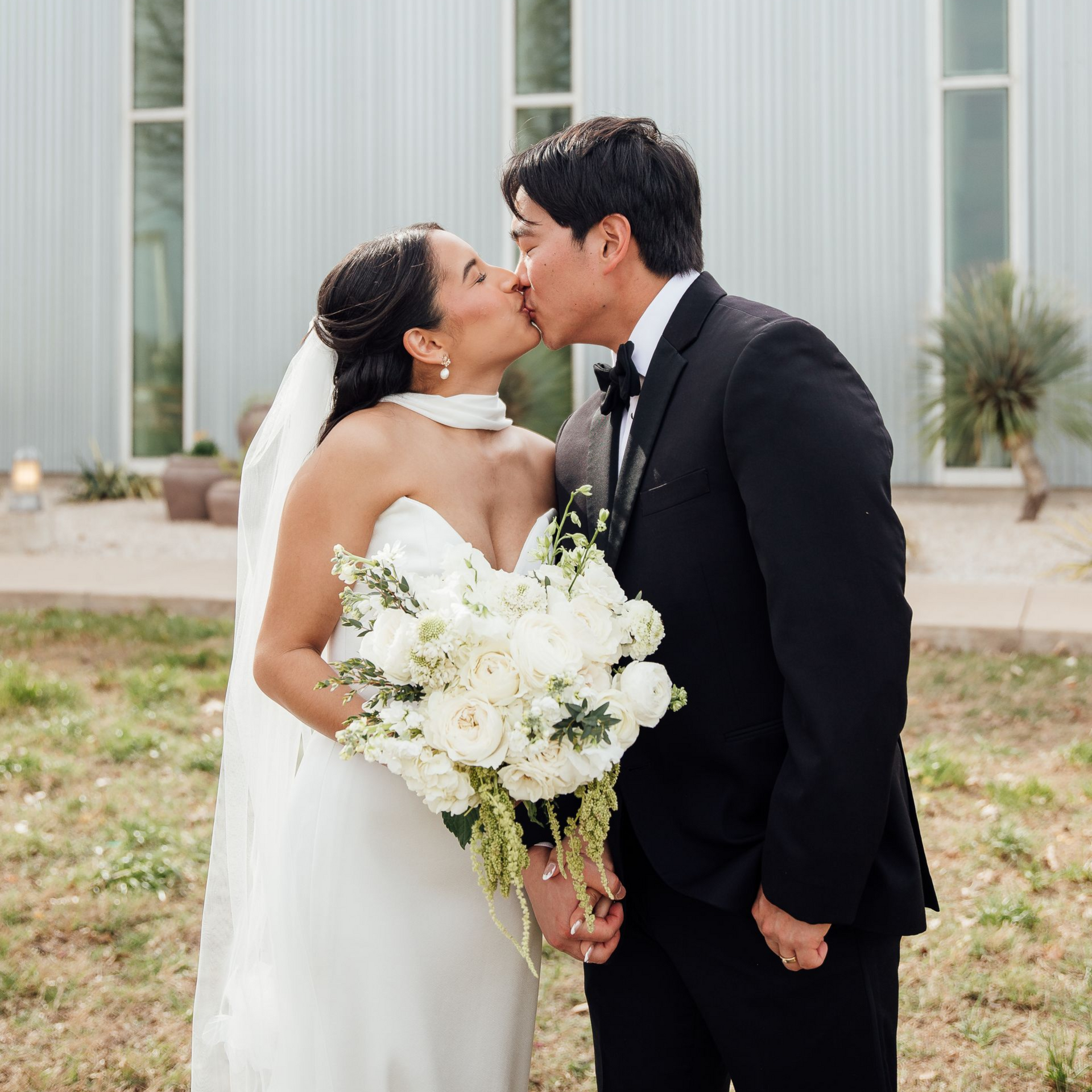 A bride in a white wedding gown and veil kisses a groom in a black tuxedo while holding a large white floral bouquet.
