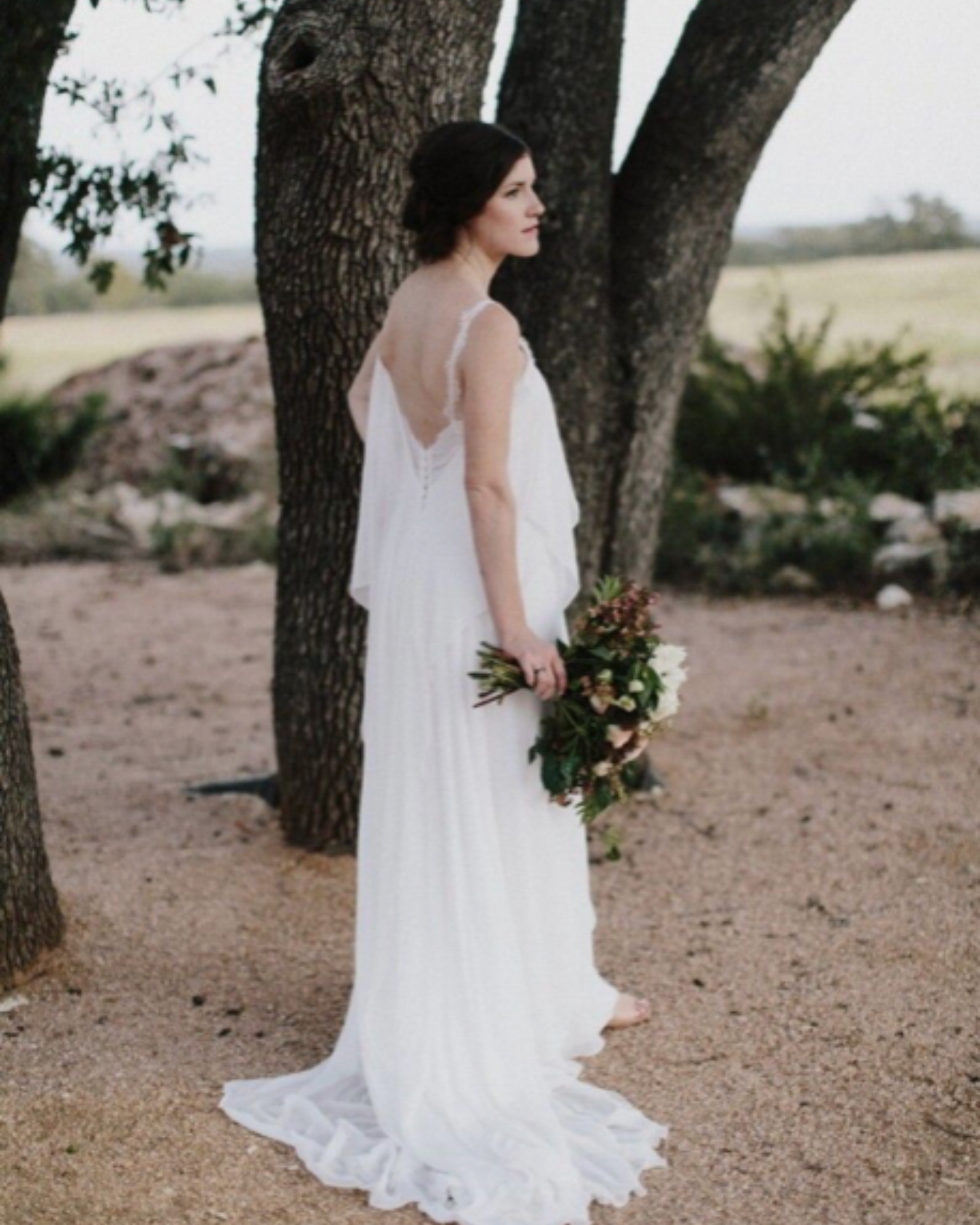 A bride in a white dress is standing next to a tree holding a bouquet of flowers