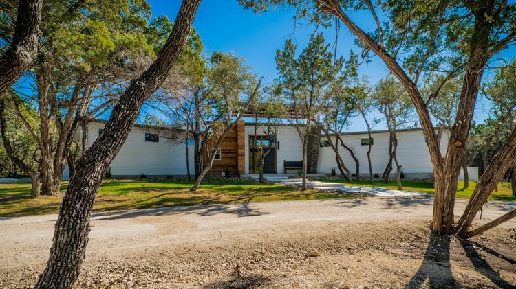 A white single cottage that is surrounded by trees and a dirt road. This is a boutique style hotel. 