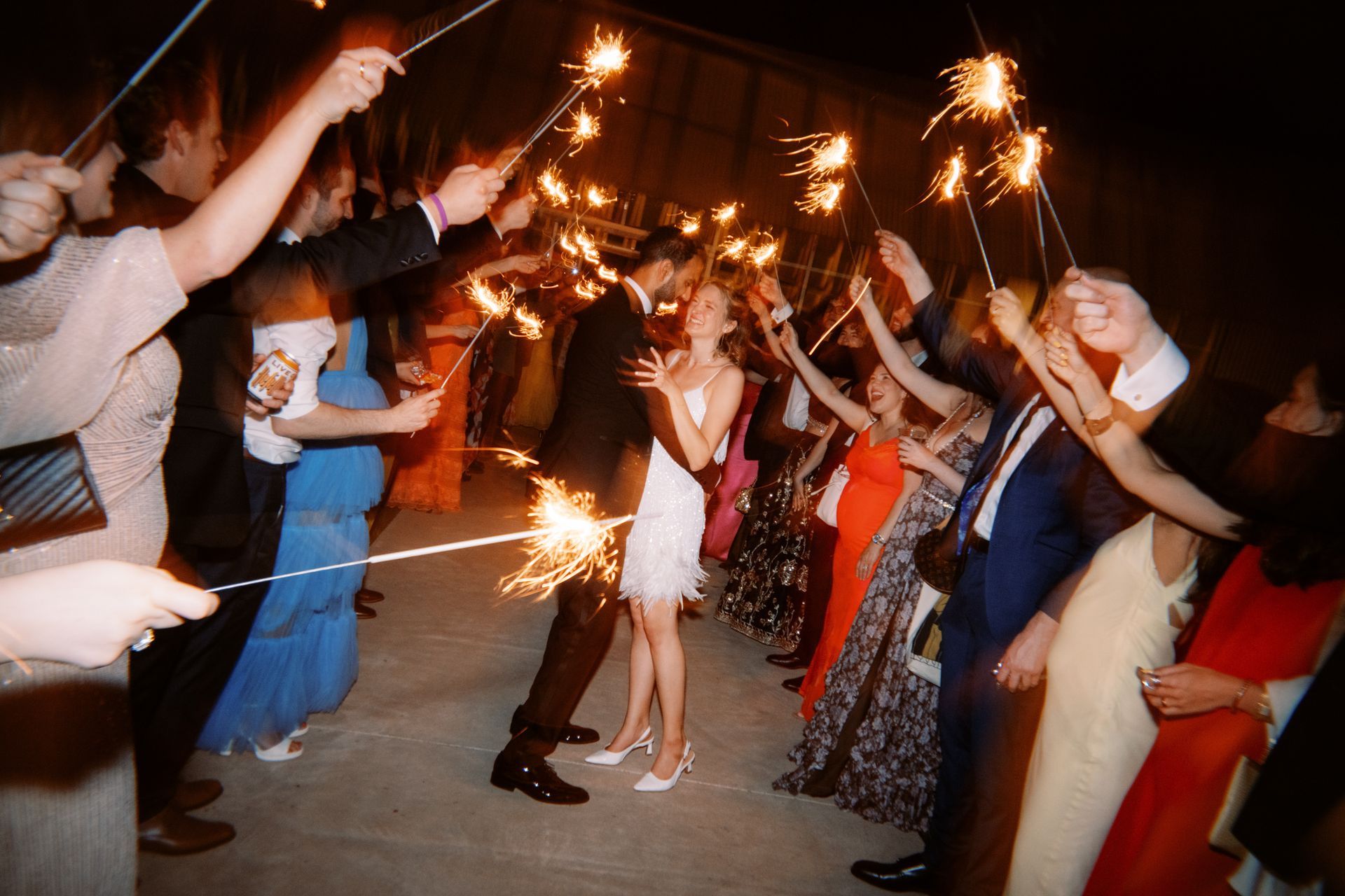 A couple dances in the center of a crowd holding lit sparklers at night, creating a festive, glowing send-off atmosphere.