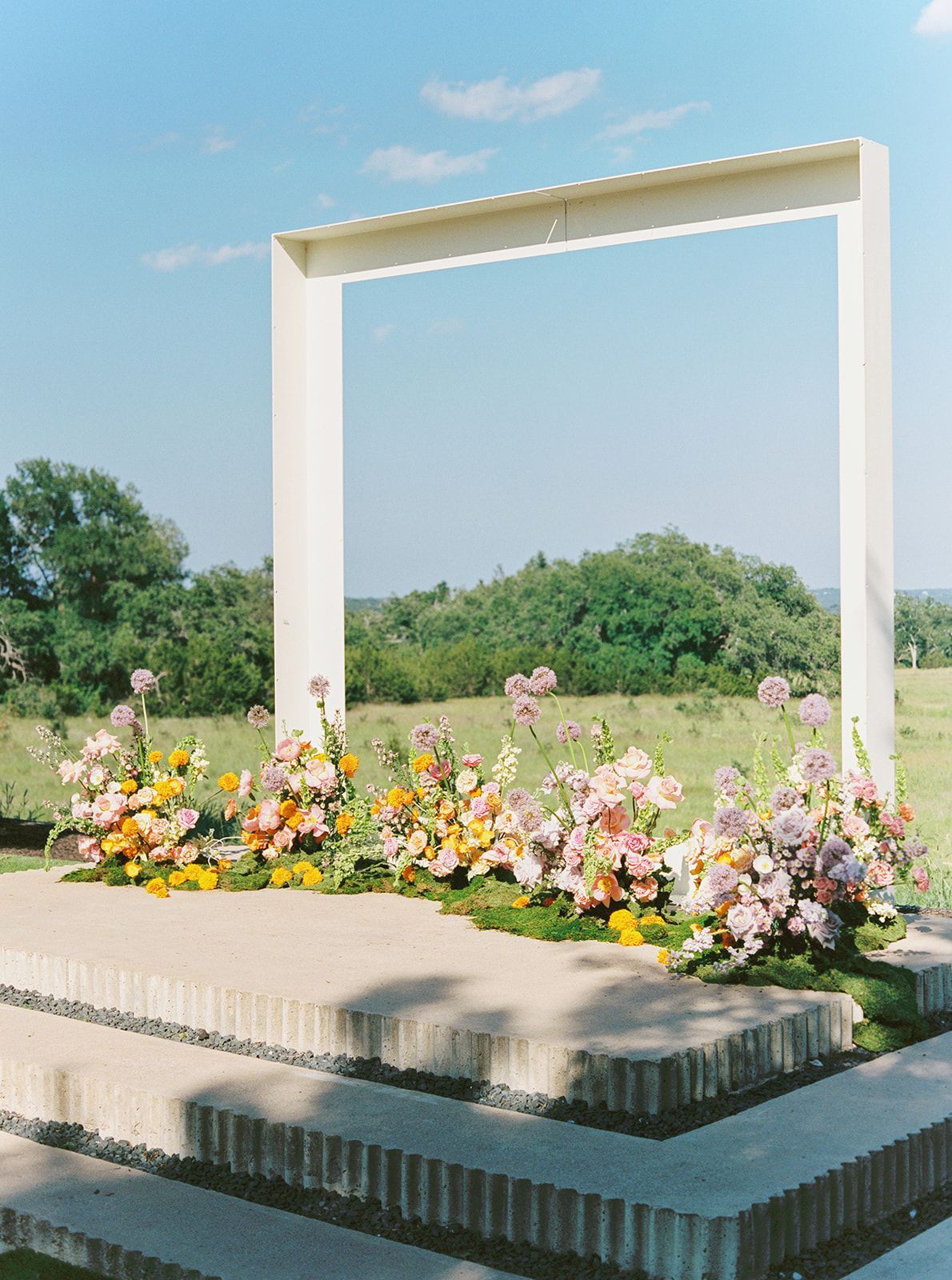 A rectangular white frame stands on tiered concrete steps outdoors, adorned with clusters of pink and yellow flowers.