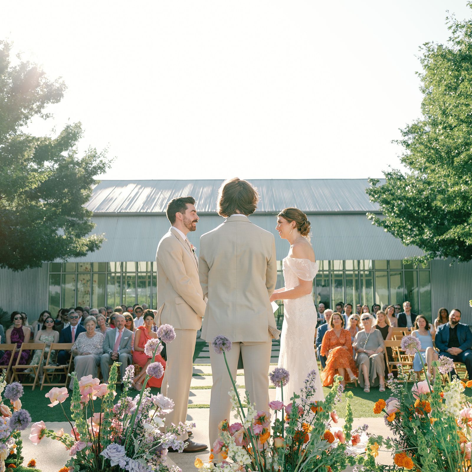 A couple stands at an outdoor wedding ceremony, facing an officiant before guests seated in a sunlit, garden-like setting.