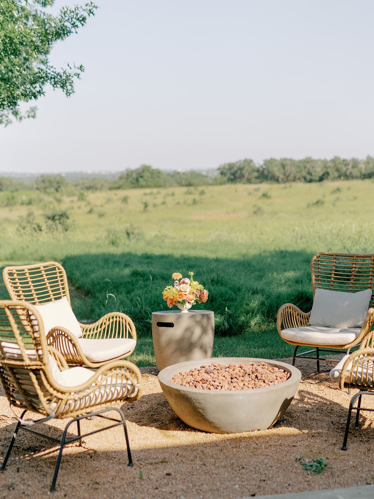 Rattan chairs surround a concrete fire pit on a gravel patio overlooking a grassy field under a bright, clear sky.