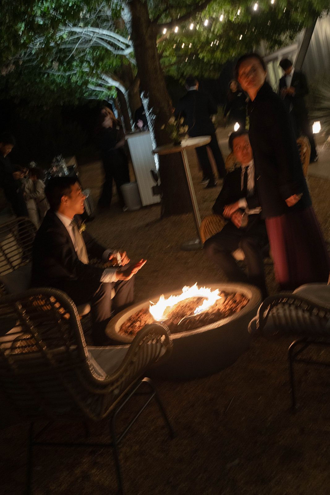 People sitting around a circular outdoor fire pit at night, illuminated by string lights and the flames.