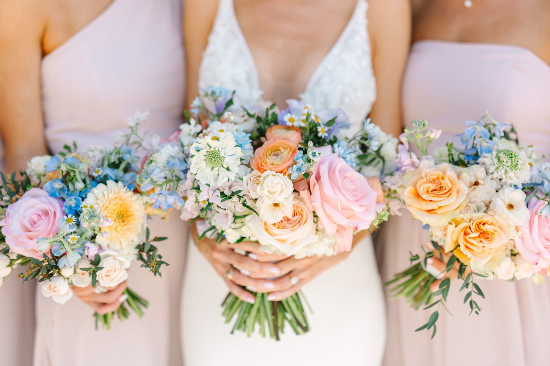 A bride and two bridesmaids in blush dresses hold floral bouquets with roses and blue flowers in a soft, bright setting.