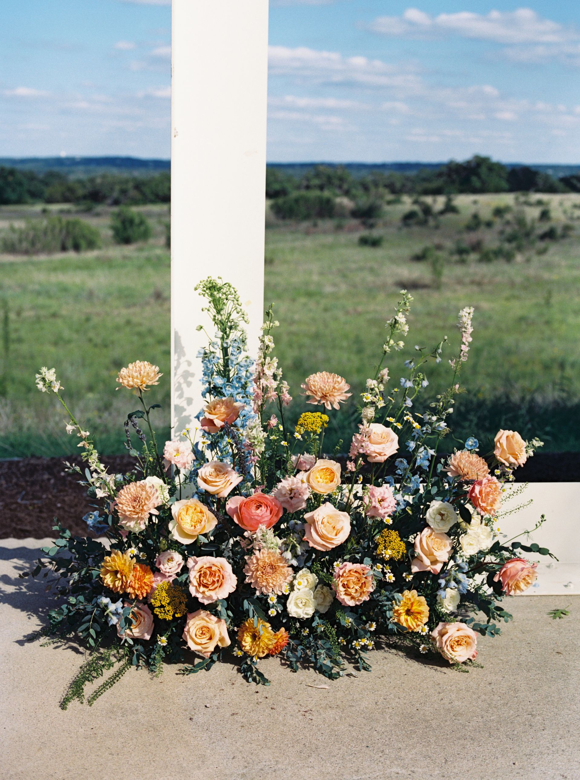 A lush, colorful floral arrangement of peach roses, orange blossoms, and blue delphiniums sits by a white post outdoors at Prospect House.