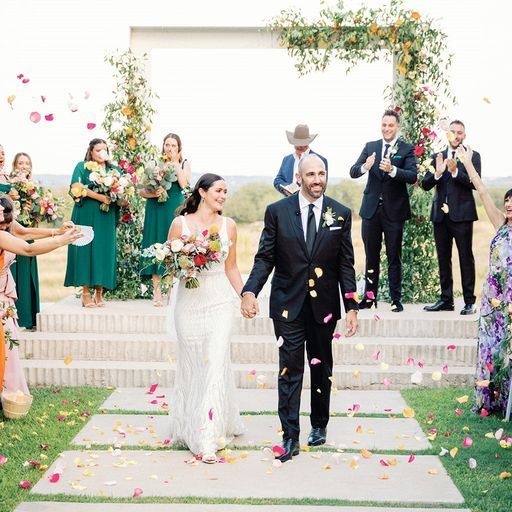 A bride and groom walk hand-in-hand down a path as guests toss flower petals, with a wedding party in the background.