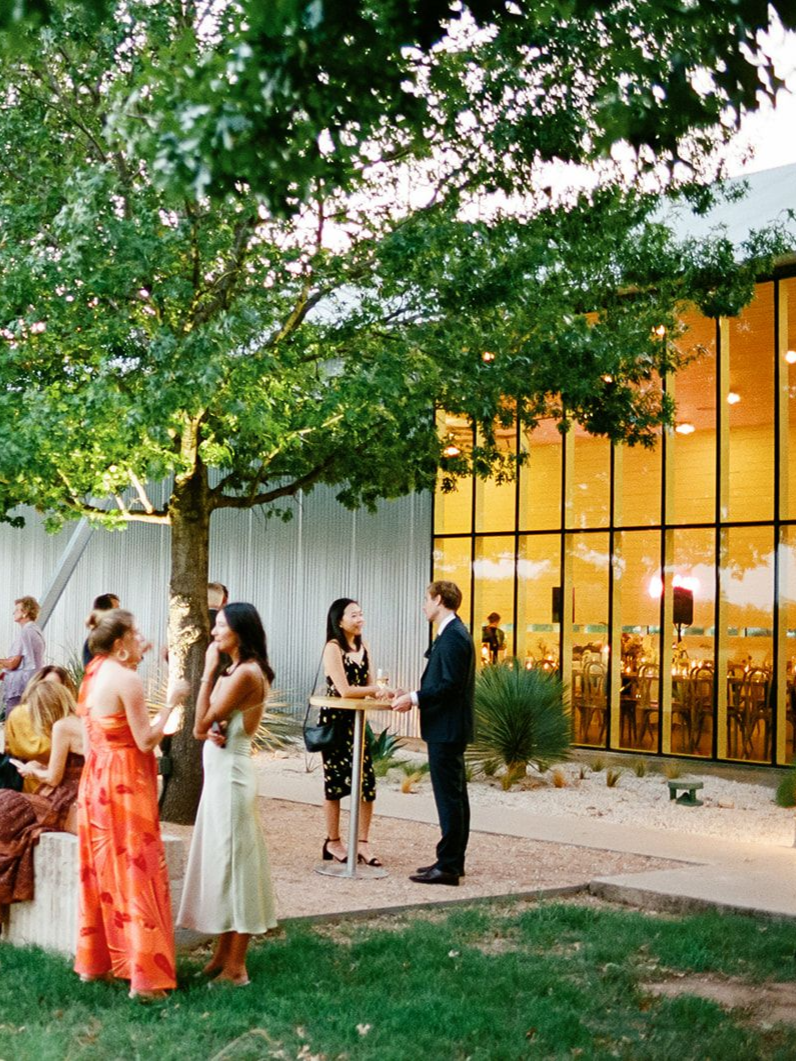 People in formal attire converse outdoors near a large, glowing glass building during an evening event under trees.