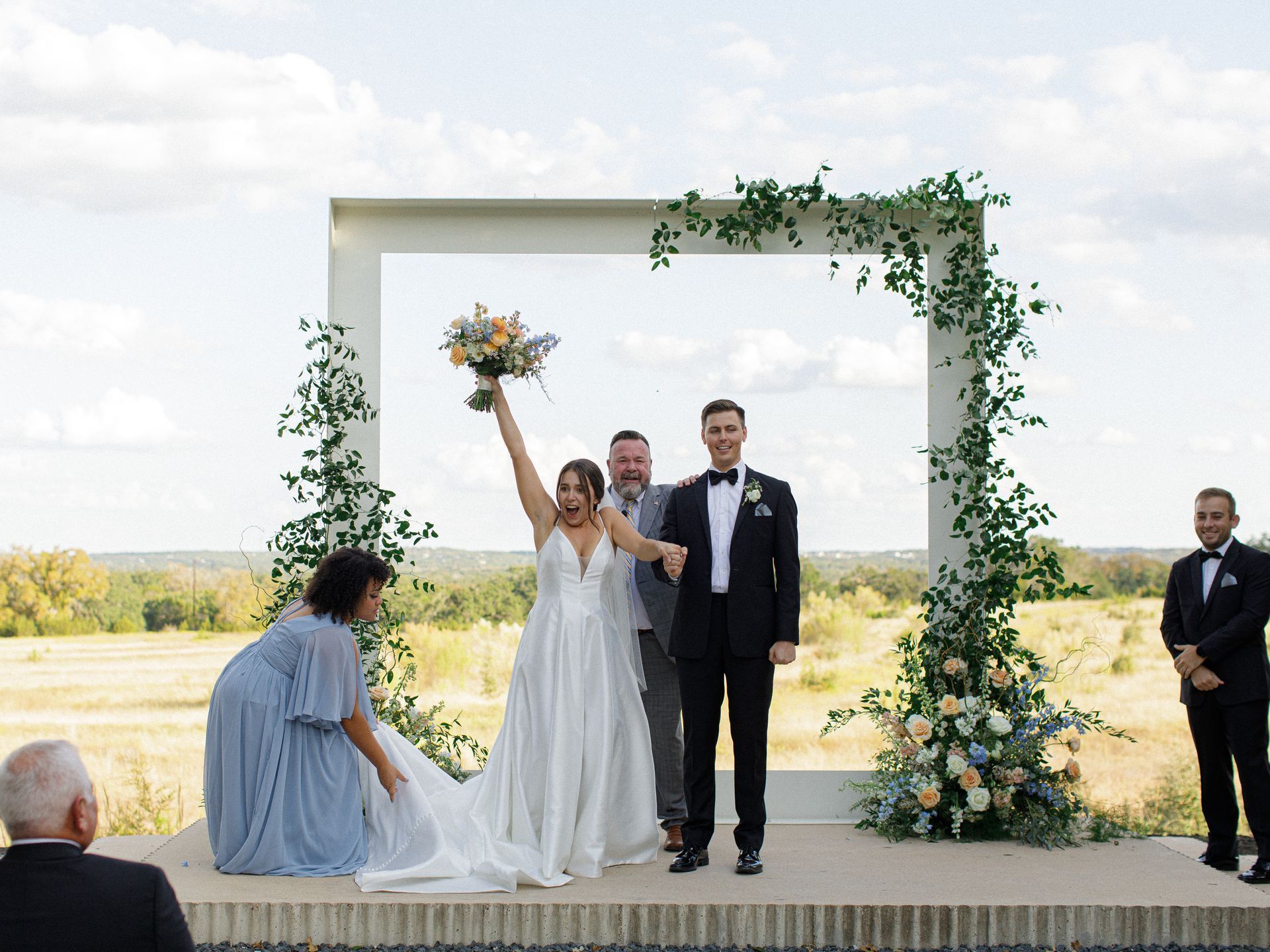 A bride cheers with a bouquet while standing with a groom and officiant under a floral arch during an outdoor wedding at Prospect House. 