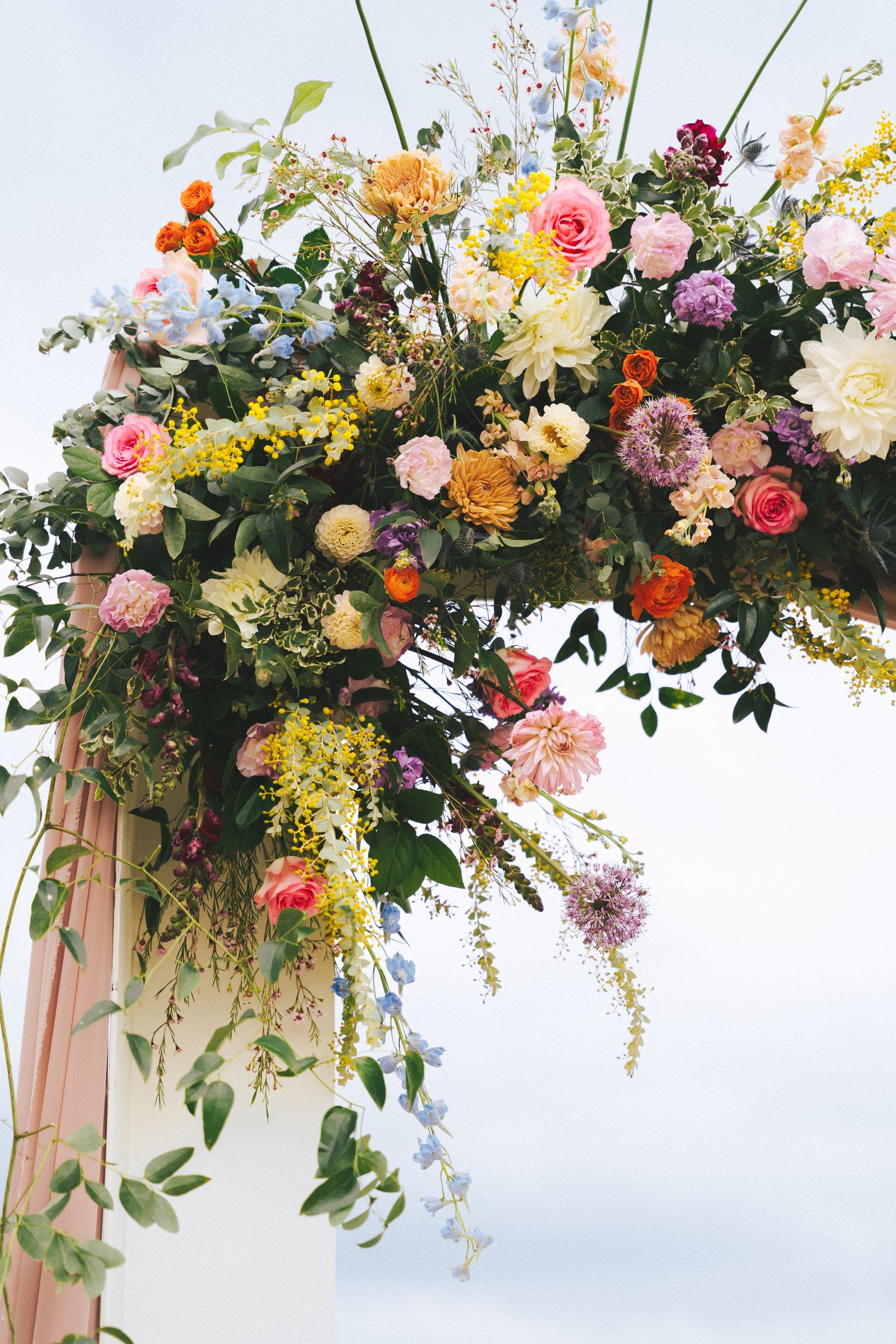 A vibrant, multi-colored floral arrangement with roses, dahlias, and greenery decorating a wedding arch against a sky.