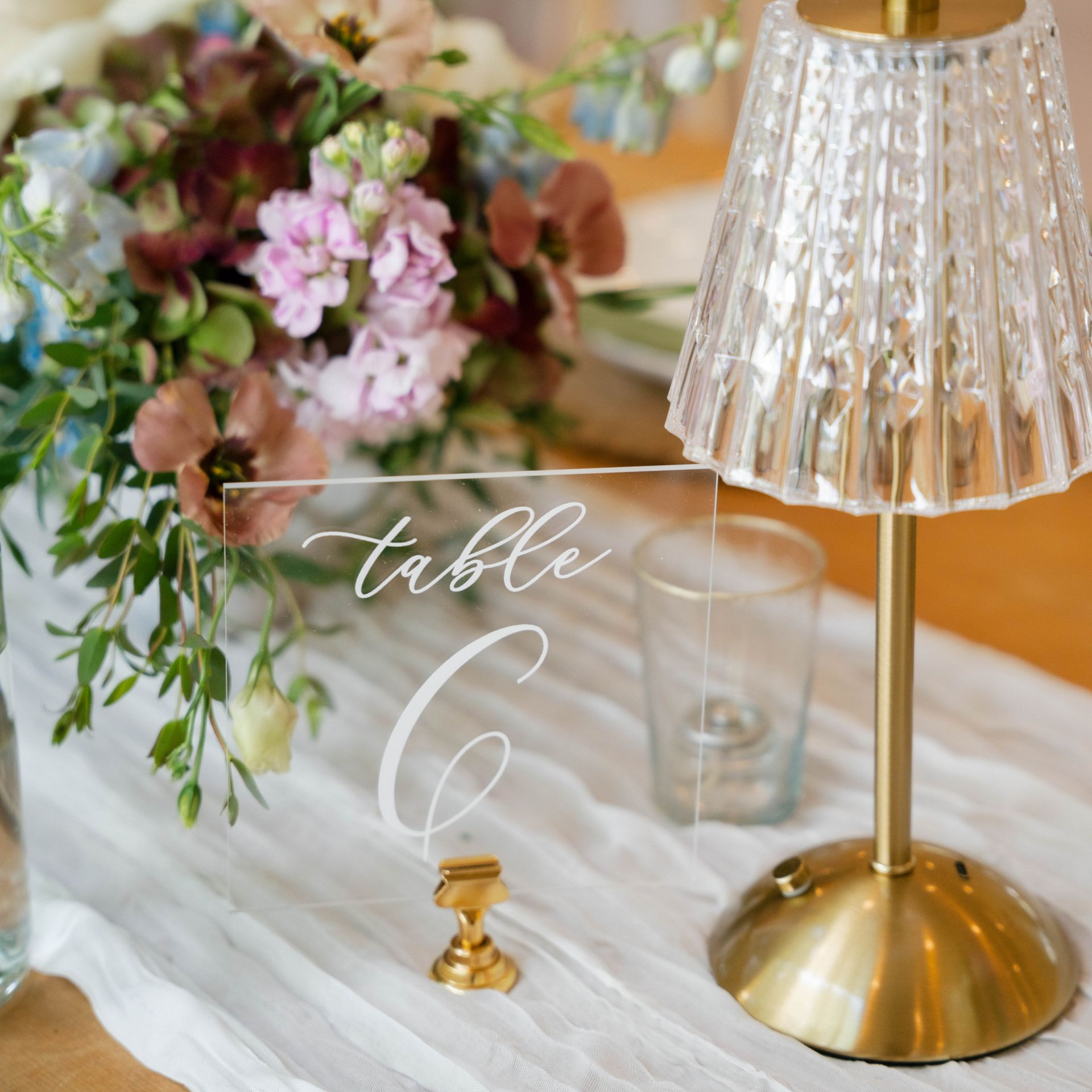 A clear acrylic table number six sign next to a gold-base lamp and a bouquet of flowers on a white runner.