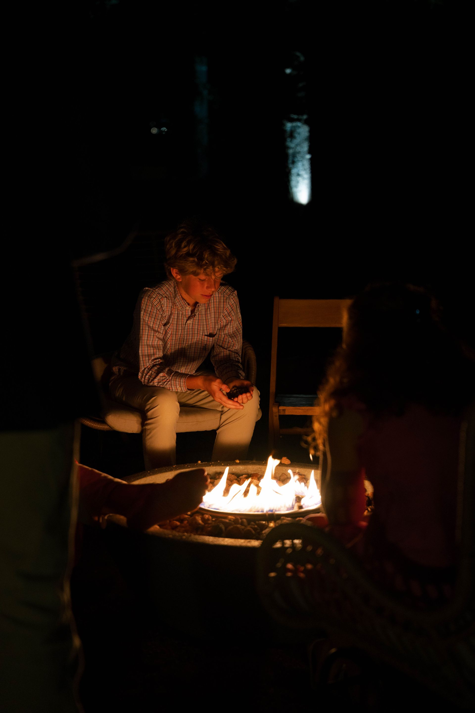 People sit around a fire pit at night, with the flames casting a warm glow on them in the otherwise dark setting.