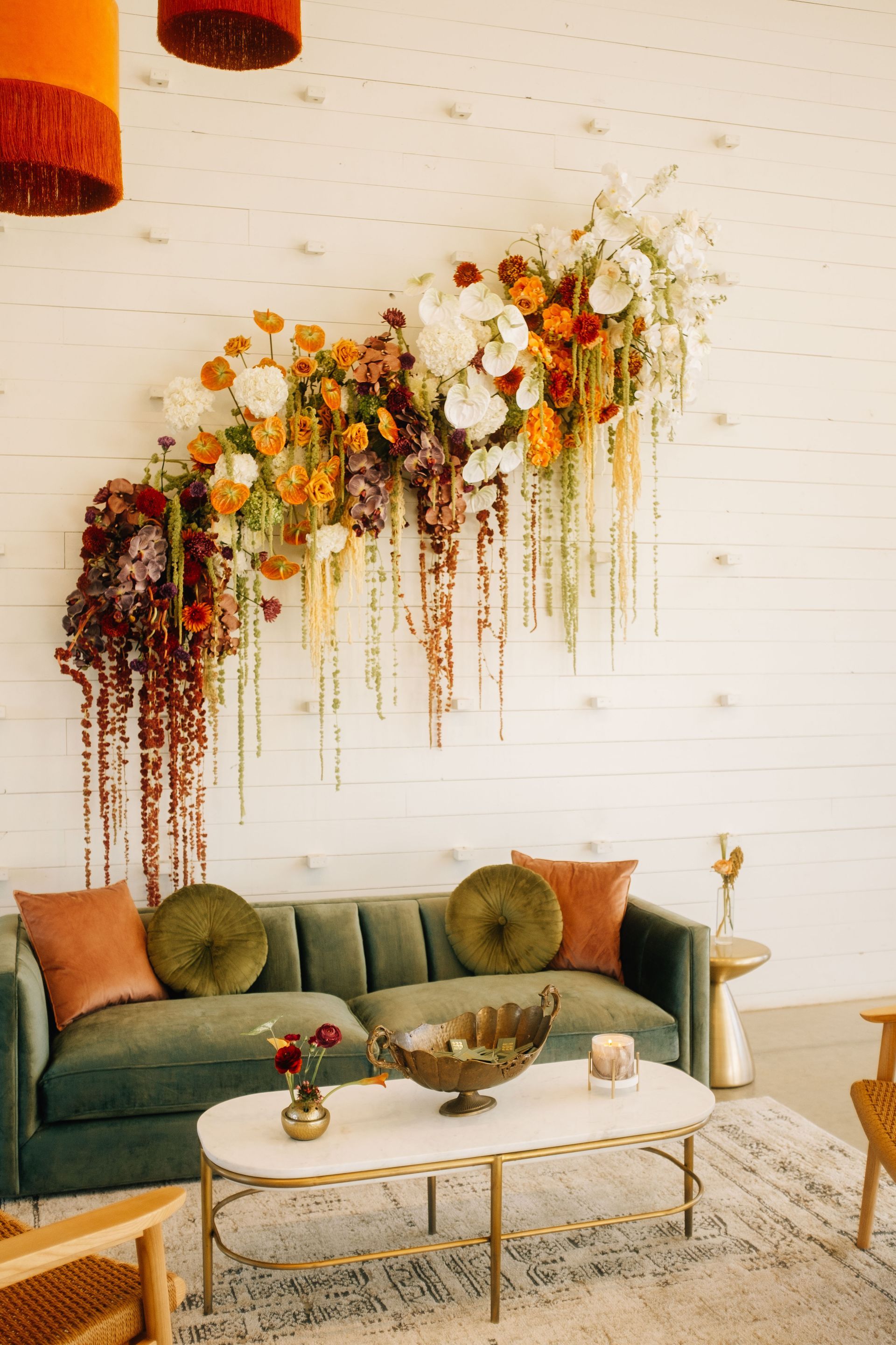 A green velvet sofa sits before a white wall decorated with a hanging floral arrangement of orange, white, and red flowers.