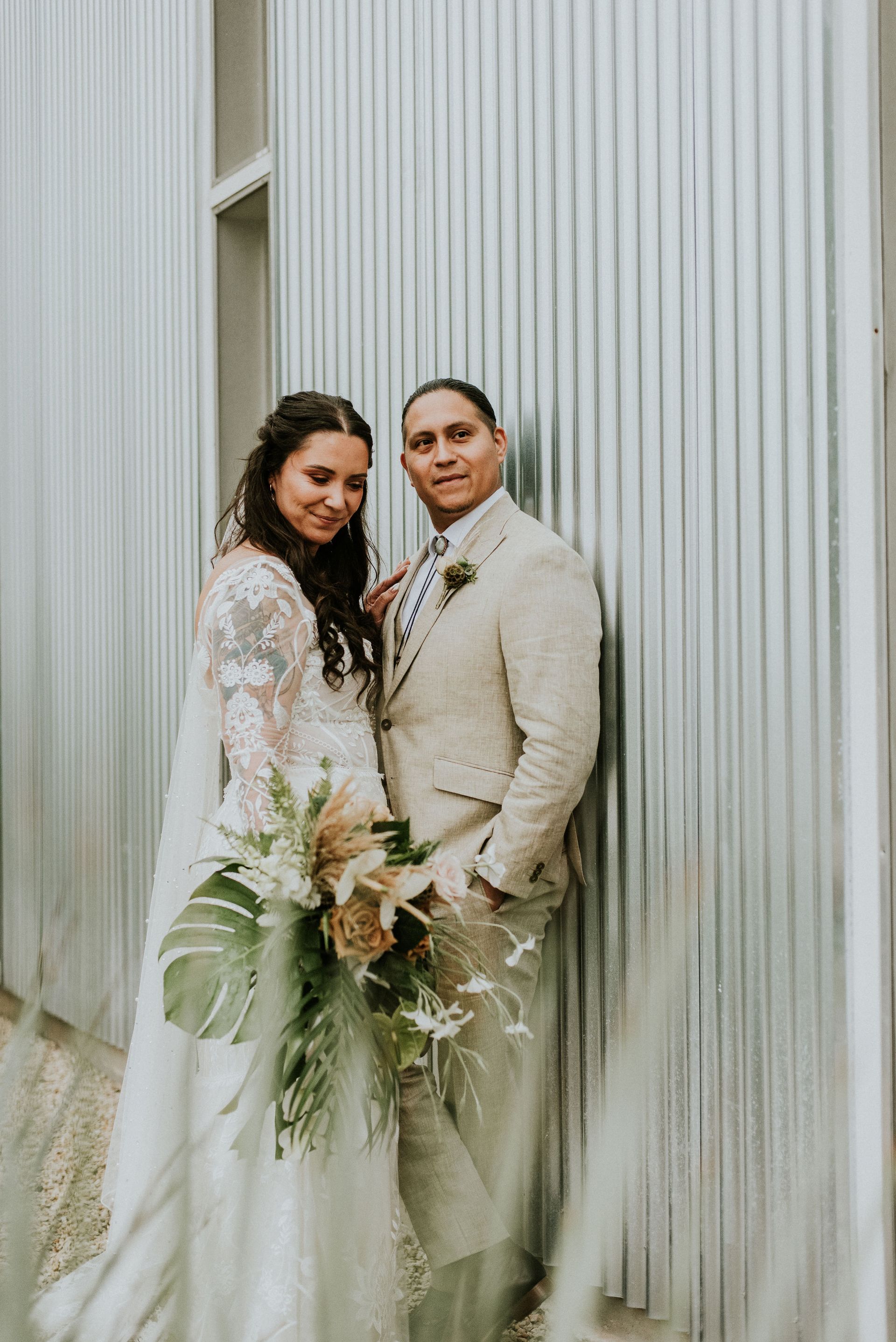 A couple stands side-by-side, dressed in wedding attire, holding a large bouquet against Prospect House's  vertical silver textured wall. 