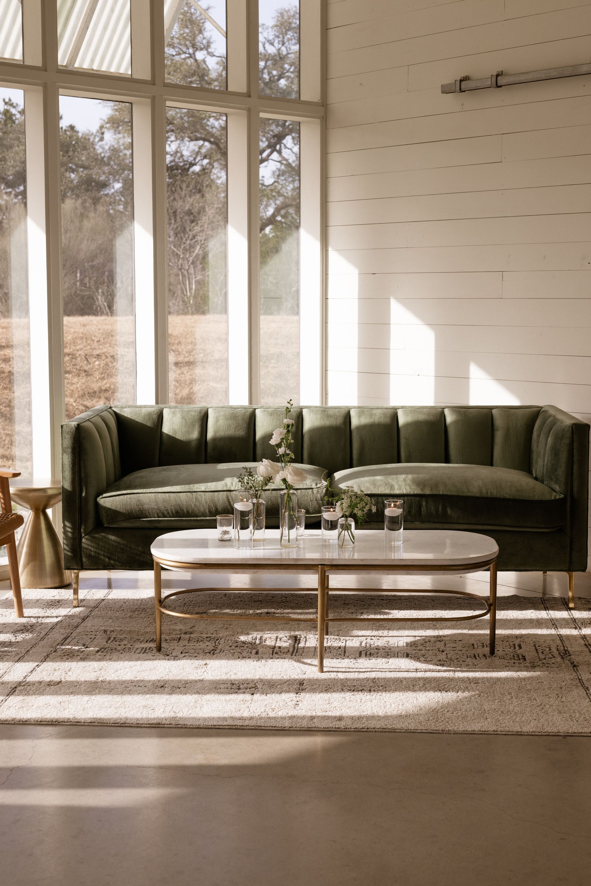 A velvet, tufted olive green sofa with a white marble coffee table sits on a textured rug in a bright, sunlit room.